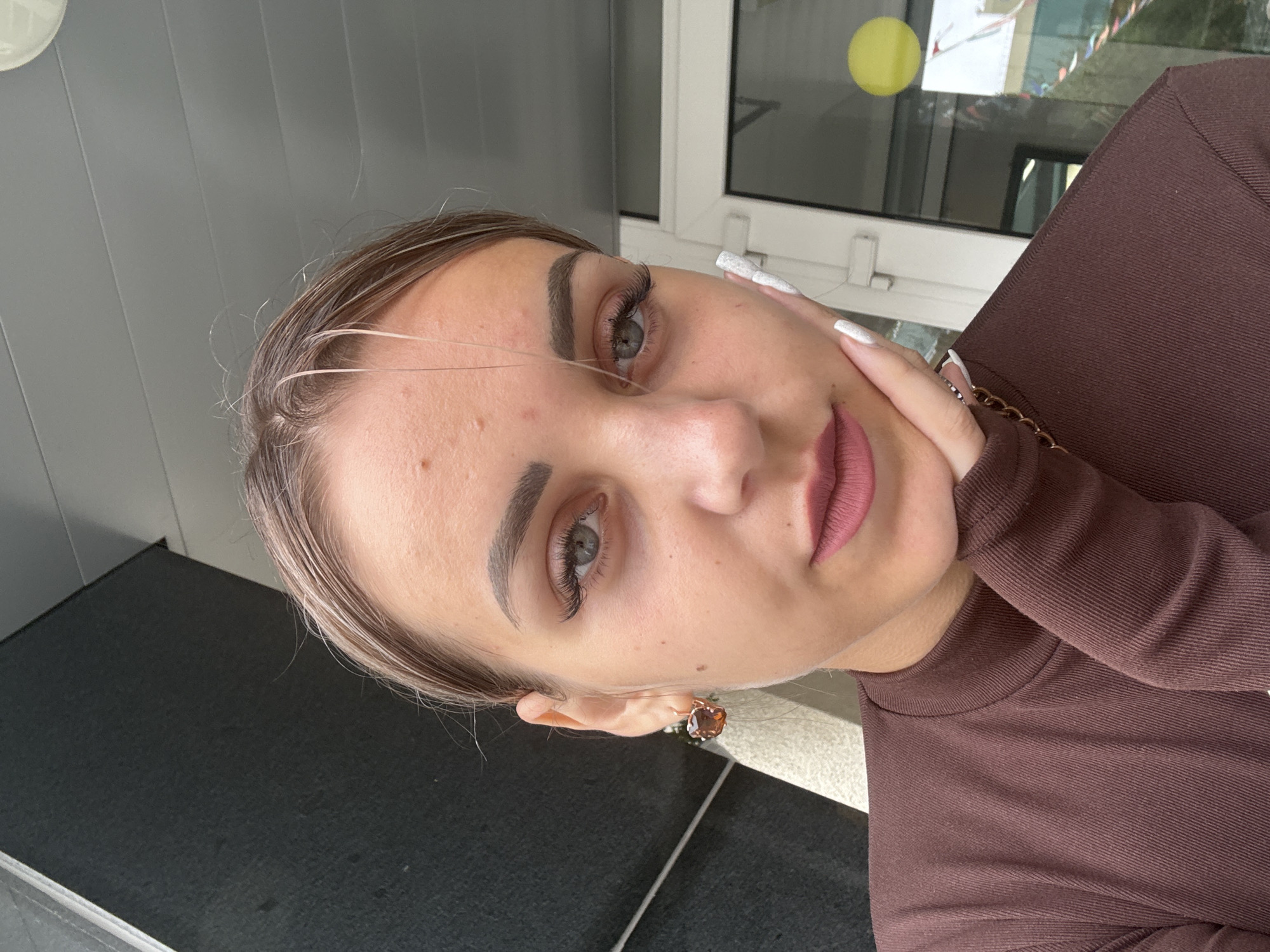 A young woman with light brown hair pulled back is shown in a close-up selfie. She is wearing a dark brown long-sleeved shirt with a high neckline. Her left hand is propped on her chin, with her manicured nails visible. She has full lips with matte pink lipstick and is wearing a large amber earring in her left ear. Her right eye is visible, with well-defined eyebrows and long false eyelashes. The background appears to be outdoors, possibly on a porch or patio, with a dark gray wall to the left and a building with windows and a yellow circle sticker in the background. The image does not contain information to determine the location in Yuzhno-Sakhalinsk, Russia.