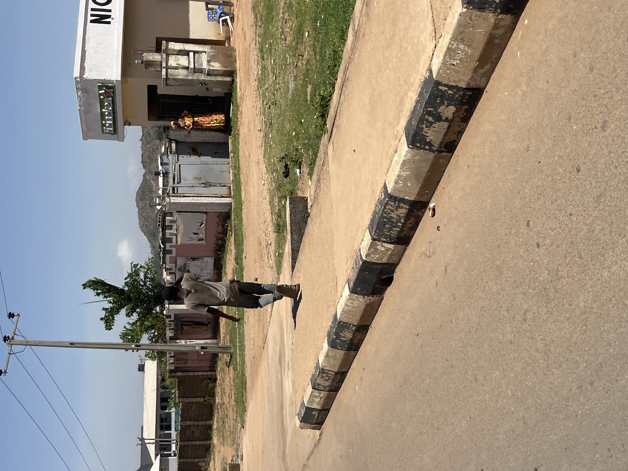 The image depicts an outdoor scene in Gura Top, Nigeria. A man wearing light-colored clothes and sandals is walking along a sidewalk with black and white striped curbing. The road is to his right. Adjacent to the sidewalk is a grassy area with sparse vegetation. Buildings of various construction styles are visible in the background, including a structure with the word "NIGER" visible on its facade, next to the inscription "Fibertex". Another person is wearing a colorful robe and stands outside one of the buildings. In the distance, a small mountain is visible. A utility pole with electrical wires stands near the left side of the image.