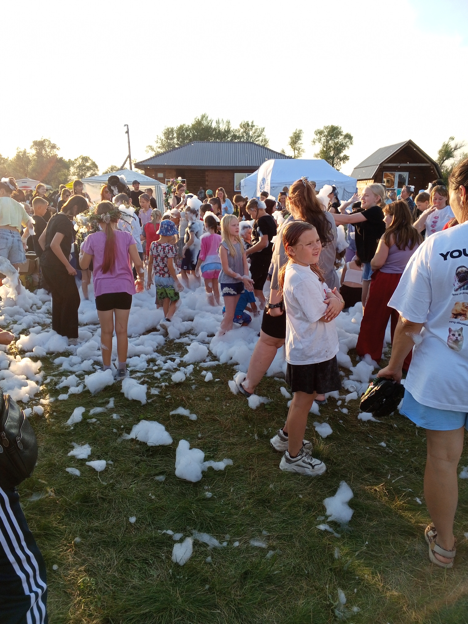 In an outdoor grassy area with scattered white foam, a large group of people, predominantly children and young adults, are gathered at what appears to be a daytime event. The scene is populated with individuals of various ages and genders, dressed in casual summer attire. Some are interacting with the foam, while others are observing or conversing. In the background, two wooden structures with dark roofs and a white tent-like structure are visible, suggesting a fair, festival, or organized activity. The sky is bright, indicating daylight, with some trees visible in the distance. The overall atmosphere seems lively and recreational.