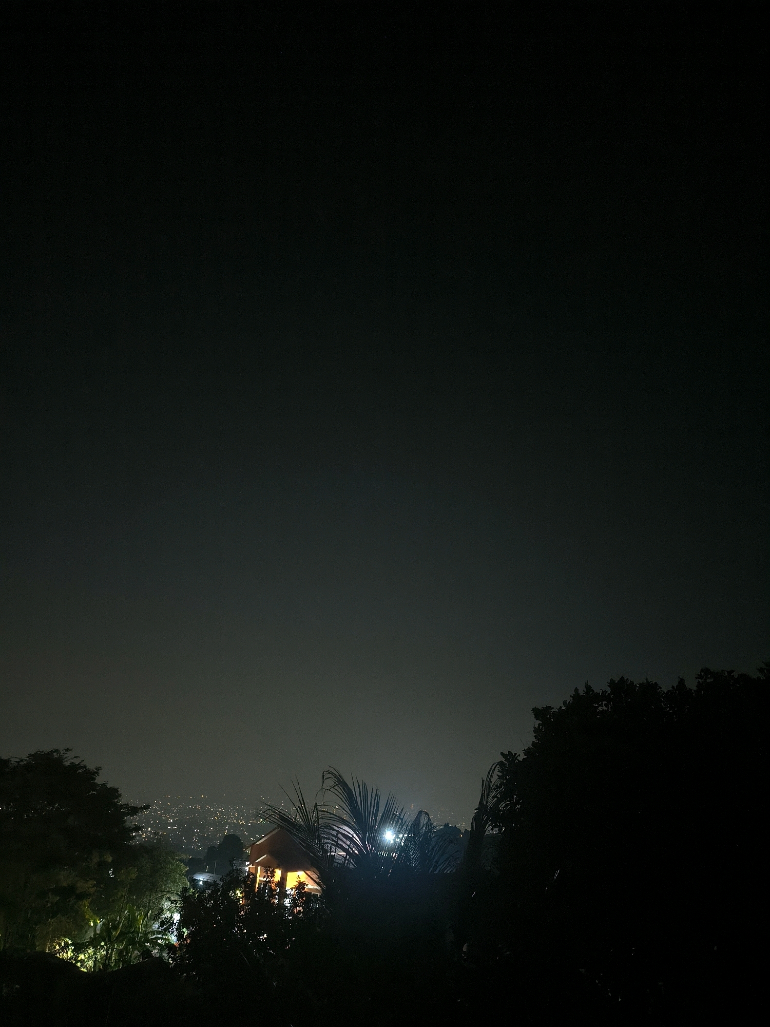This is a nighttime photograph taken from an elevated position overlooking the city of Cilengkrang, Indonesia. The foreground is dominated by dark, silhouetted trees and foliage, with some artificial lights visible amongst the greenery. In the mid-ground, a small, orange-lit structure, possibly a house or a pavilion, is visible, with palm fronds and other vegetation around it. Beyond this, the city sprawls out, characterized by a vast expanse of small, scattered lights indicating buildings and streetlights. The sky is dark and hazy, with a faint glow from the city lights. The overall scene conveys a sense of a quiet, residential area at night, with the distant city lights creating a soft backdrop.