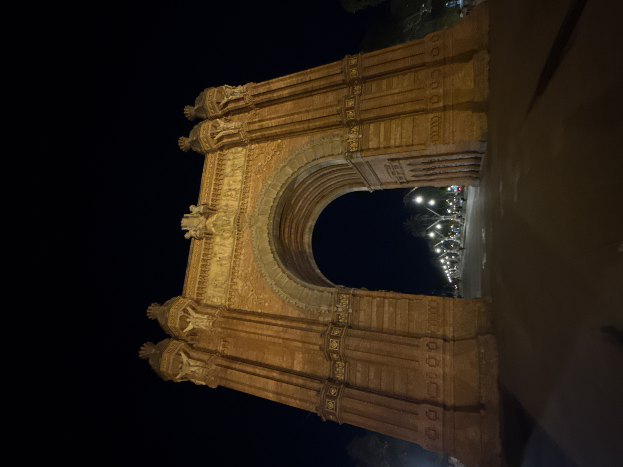 The image shows the Arc de Triomf in Barcelona, Spain, at night. The triumphal arch is made of red brick and has intricate carvings. It is lit up by lights, and the street is visible through the arch with lamp posts. The sky is dark.