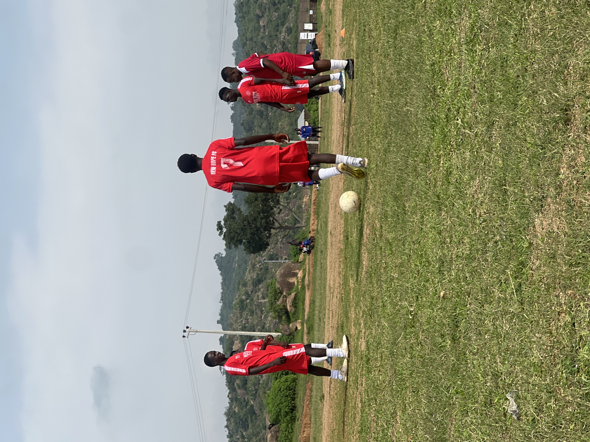 In Jos, Nigeria, on a sparsely vegetated, dry football pitch, a group of young men in red football kits are practicing. The sky is overcast with light clouds. The background features rocky hills and some trees. A football lies on the grass near the foreground, and one of the players in the center is facing away from the camera, his jersey displaying the number 7 and the words "NEW HOPE FC." Two other players in similar kits are standing nearby, looking towards the center. Another player on the left is also in a red kit. In the far background, several people are seated in the shade of a structure, and a few figures are standing near what appears to be a building.