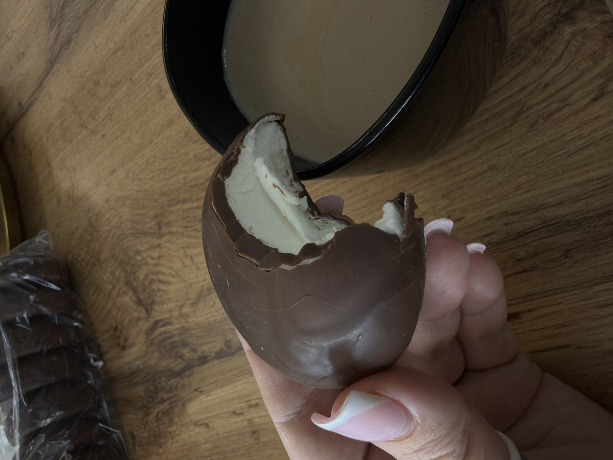 A close-up shot depicts a person holding a partially eaten chocolate egg. The egg has a thick chocolate shell and a white, creamy filling. Two bites have been taken out, revealing the filling. The person holding the egg has pink and white manicured nails. Behind the egg, there is a black bowl containing a light tan-colored liquid. To the left of the egg and bowl, there is a partially visible package of chocolate biscuits. The background is a wood-grain surface. The image's location is unknown. The city of Chaikovsky, Russia, is not identifiable in the photo.
