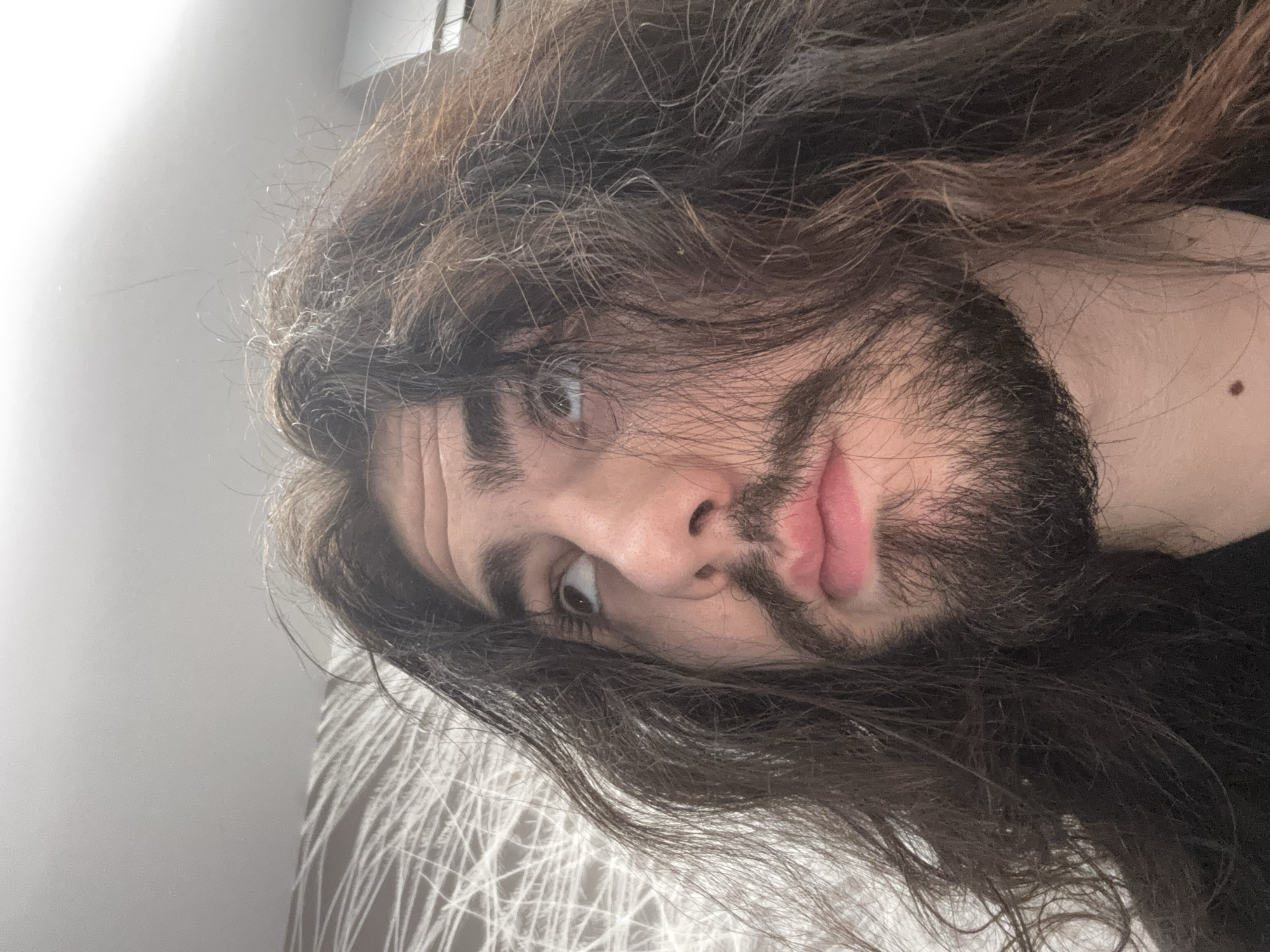 A close-up, medium shot captures the face of a person with long, dark, wavy hair, a mustache, and a beard. The person is looking upward and to the left, with their head tilted back slightly. Their lips are parted, revealing a hint of their teeth. The background consists of a textured wall with a white, abstract pattern and a plain gray ceiling. The lighting appears to be artificial, casting a slight shadow on the person's face. The overall impression is of a casual, informal setting, likely indoors. There is no identifiable location in the city of Unknown, Unknown.