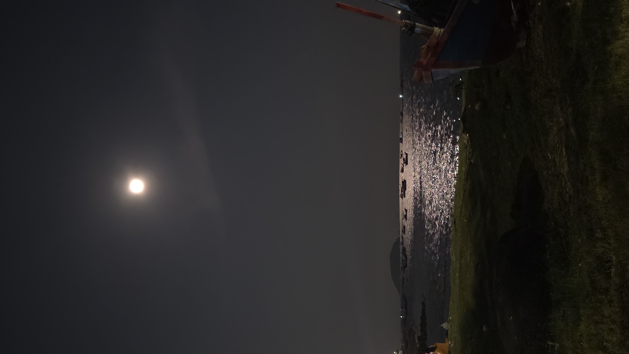 At night in Bắc Nha Trang, Vietnam, a boat rests on a grassy shore beside the ocean. Moonlight illuminates the water, creating a shimmering effect. The sky is dark, with a bright moon dominating the left side of the frame. Several smaller boats are visible in the distance on the water.