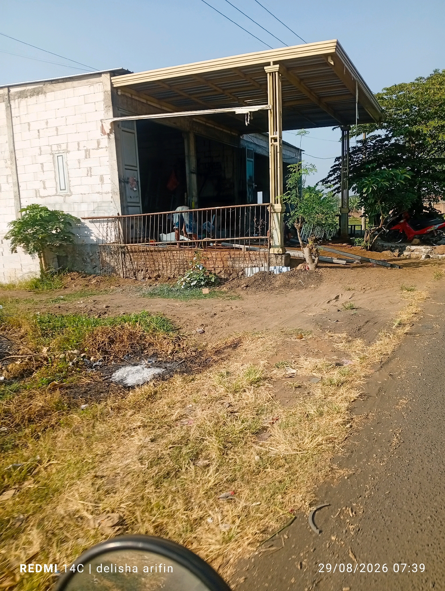 The image captures a street-level view of a building with a corrugated metal roof supported by beige posts, located in Lebaksari, Indonesia. The building's facade is constructed from white brick, and it features a large, open entrance revealing the interior. A metal railing runs in front of the building, and in the foreground, dry grass and dirt cover the ground. To the right, a paved road is visible, and beyond that, a lush green tree. The scene is illuminated by clear daylight, indicating it was taken during the daytime. A timestamp in the lower right corner reads "29/08/2026 07:39", and a watermark at the bottom left states "REDMI 14C | delisha arifin".