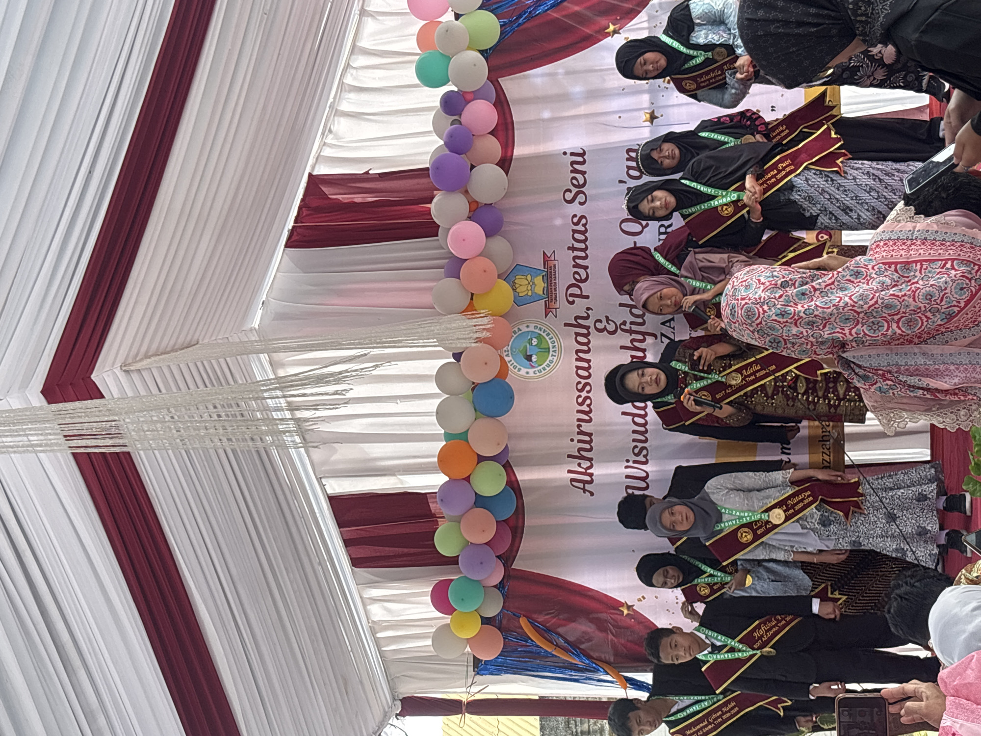 The image depicts an outdoor awards ceremony held under a white and maroon tent. A group of ten individuals, mostly young women and a few young men, are standing on a stage, some wearing sashes and medals. They appear to be participating in a graduation or recognition event. Behind them, a banner reads "Akhirussanah, Pentas Seni & Wisuda Hafidzah Qur'an." The stage is decorated with a string of colorful balloons and a star motif. Several people are seated in the foreground, some holding up cell phones to take pictures, indicating an audience. The overall atmosphere suggests a formal yet celebratory event. The presence of the banner and the nature of the event strongly suggests this is taking place in Sukabakti, Indonesia, given the context provided.