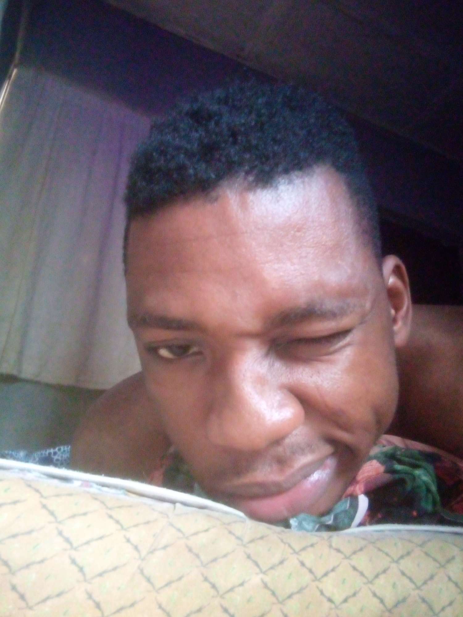 A close-up indoor shot features a man lying down, winking with his left eye. He has short, dark, textured hair and appears to be resting his head on a pillow with a patterned cover. Behind him, there is what looks like draped fabric or a curtain. The location is unknown.