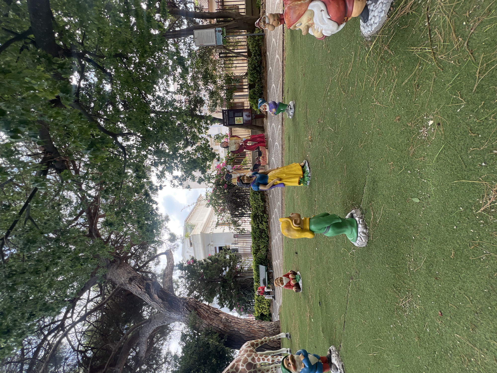 The image depicts a park in Palmi, Italy, featuring a collection of dwarf figurines and a statue of Snow White arranged on artificial turf. In the background, a giraffe statue is partially visible to the left, and behind that, a white building with ornate architectural details stands. A few other statues, including what appear to be costumed figures, are positioned further back near a fence. A person is seated on a park bench under a large tree in the middle ground. The ground is covered in artificial green grass with scattered twigs and small stones. The trees overhead cast shadows, and the sky is partly cloudy.