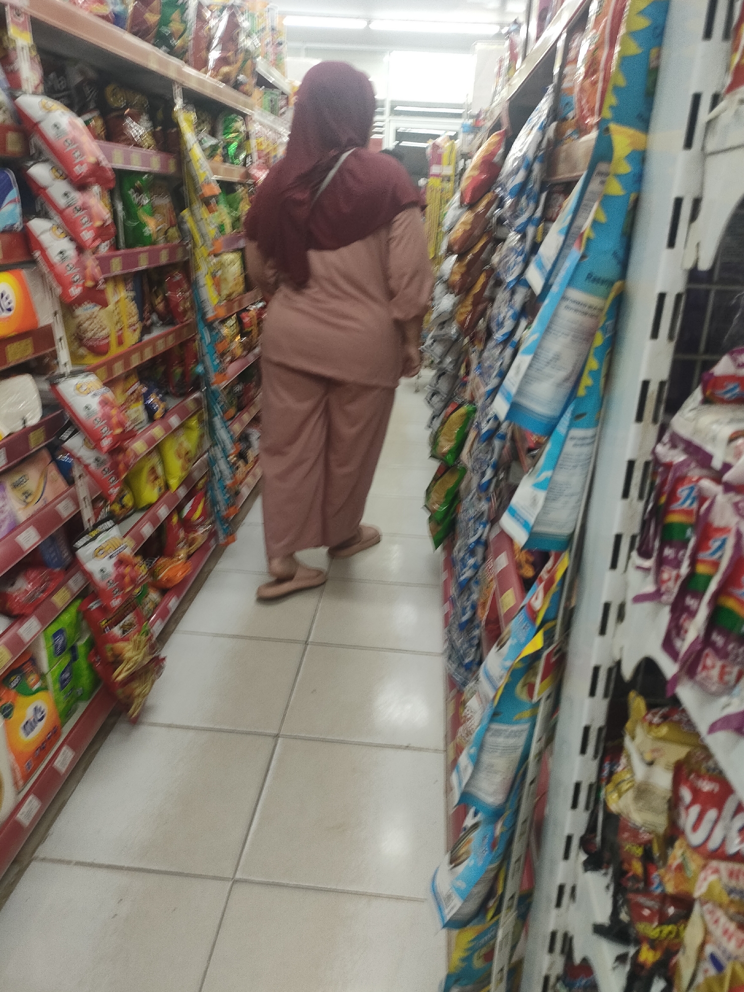The image shows the interior of a minimarket in Desa Banyumas, Indonesia. A woman, wearing a maroon hijab and a pale pink tunic and pants, is walking away from the camera down an aisle lined with shelves stocked with various packaged snacks and food items. The shelves are red, and the floor is tiled with large, light-colored squares. The aisle is narrow, and the lighting suggests an indoor, commercial setting.