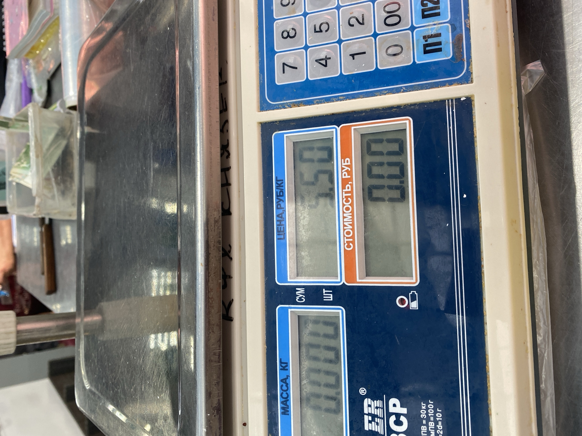 This is a close-up, overhead shot of a digital scale with a metallic platform. The scale is a commercial model with a keypad and two digital displays. The keypad has numerical buttons (0-9) and some special function keys. One display shows "5.50" and is labeled "ЦЕНА, РУБ/КГ" (Price, RUB/KG). The other display shows "0.00" and is labeled "СТОИМОСТЬ, РУБ" (Cost, RUB). Below these, another display shows "0.000" and is labeled "МАССА, КГ" (Mass, KG).
There is some text on the scale, including brand markings like "ER CP ®" and information about weight limits: "ПВ = 30 кг" (Maximum Weight = 30 kg), "МПВ=100 г" (Minimum Weight = 100 g), and "=2d=10 г" (Tolerance = 10 g).
To the left of the scale, some items are visible, including clear plastic bags containing what appear to be small items, possibly food or other goods. There's also a portion of a metallic surface that is likely part of the weighing platform or another piece of equipment. The overall setting appears to be a marketplace or a retail environment, possibly a food stall, given the nature of the items and the scale. The lighting suggests it is indoors during the day. There are no people directly visible in the frame, but the context implies a commercial transaction. The image is taken in Novaya Usman', Russia.