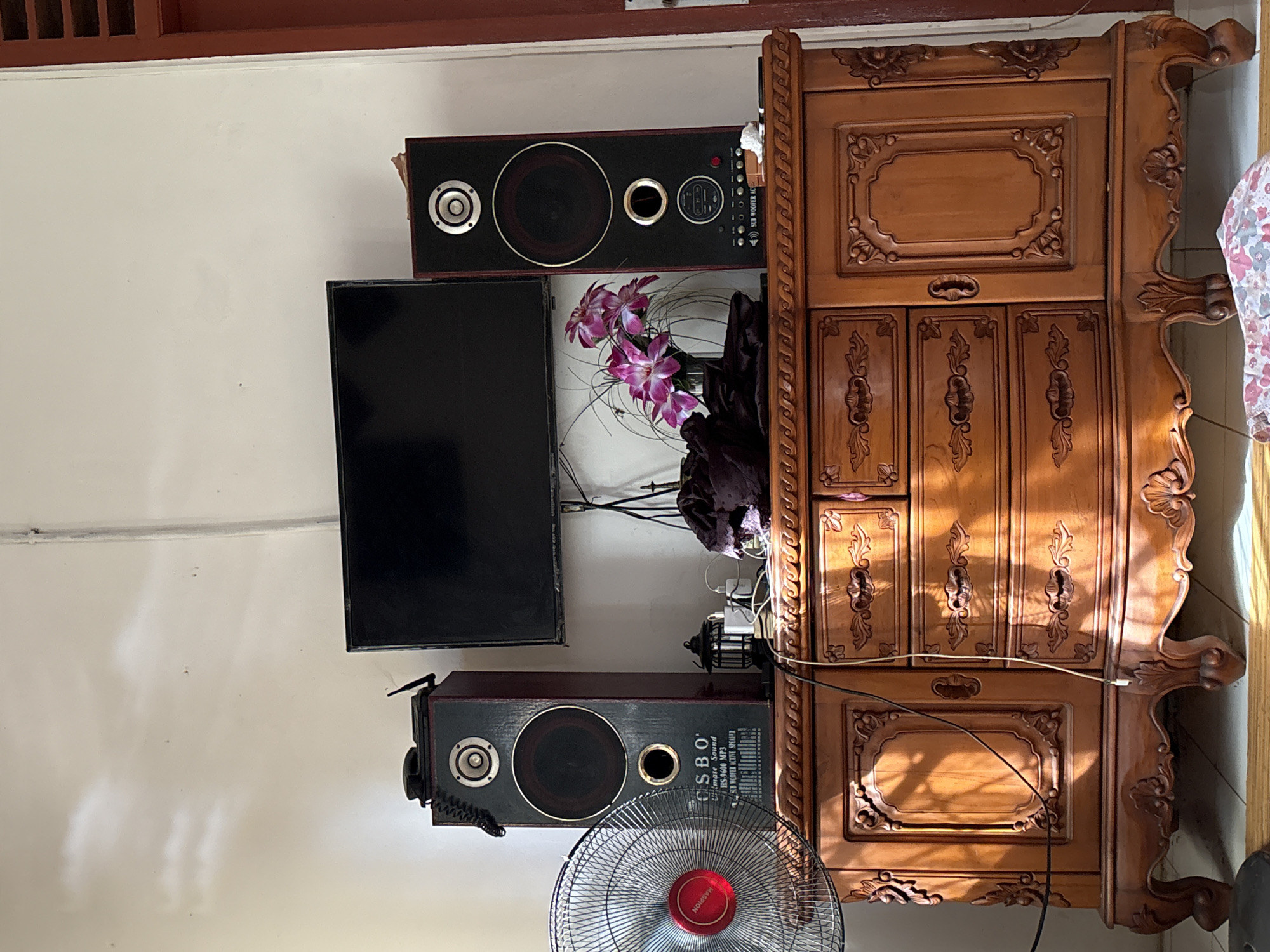 The image depicts a wooden dresser with several drawers and a cabinet, adorned with ornate carvings and elaborate legs. On top of the dresser are two tall, dark-colored speakers, a flat-screen television, a small vintage-style lantern, and a white electrical device plugged into a power strip. To the left of the dresser, a silver oscillating fan with a red center is visible. A vase with pink and white flowers stands between the speakers. The scene is illuminated by sunlight casting strong shadows across the dresser and the floor. The wall behind the dresser is painted white, with a thin vertical pipe running down it. The overall impression is of a domestic interior. The location is identified as Uleepulo, Indonesia.