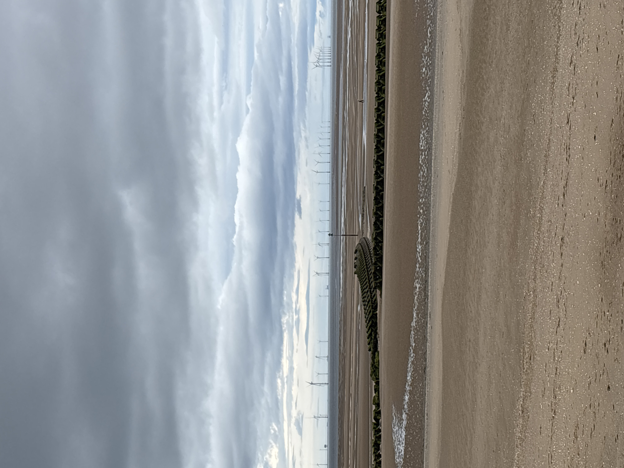 An eye-level, wide shot captures a vast, grey, cloudy sky dominating the upper half of the frame. Below, the horizon showcases an offshore wind farm, with numerous white turbines dotting the calm sea. The foreground is a wide, sandy beach, marked by the receding tide. A line of dark, textured breakwaters runs across the mid-ground, curving slightly to the right. A single person is visible in the distance, wading in shallow water on the beach.