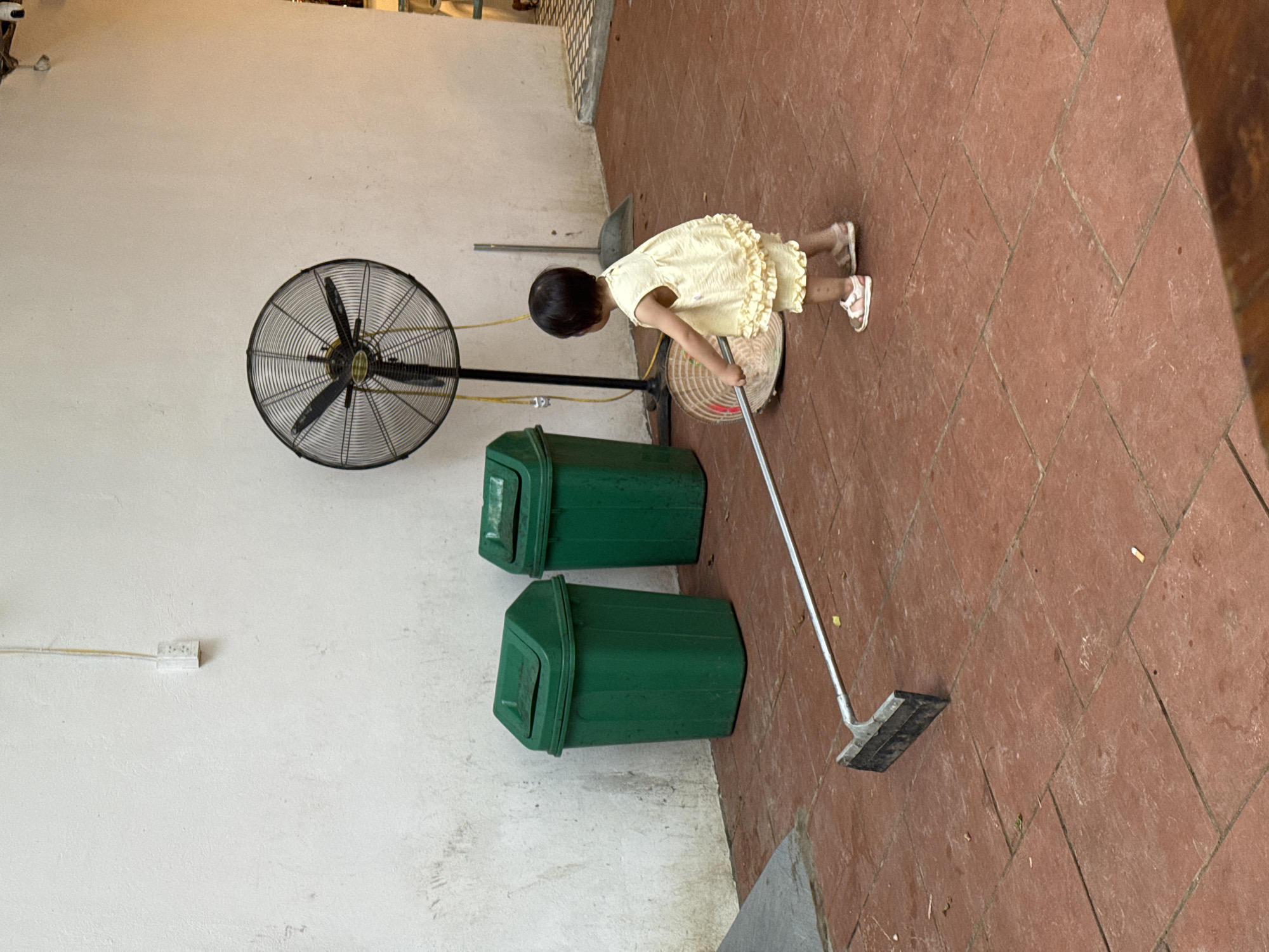 In Cổ Lũng Commune, Vietnam, a young child wearing a yellow ruffled dress and sandals is seen in an outdoor patio area. The child is holding a long-handled garden hoe, appearing to use it. In the background, two green trash cans stand against a white wall. A large industrial fan is positioned behind the child. The ground is paved with reddish-brown tiles.