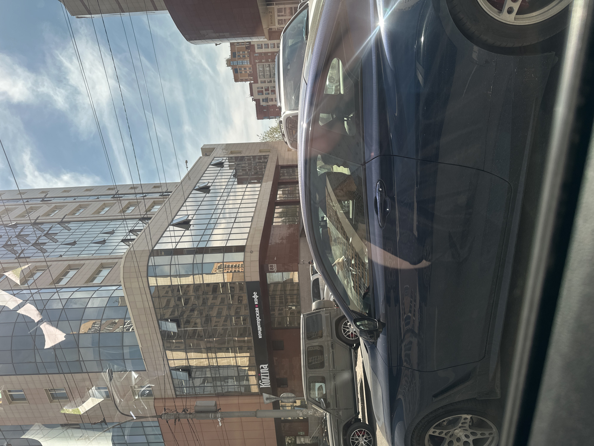 The image captures an urban street scene on a bright, sunny day, appearing to be taken from the interior of a vehicle looking out a side window. The photo is rotated 90 degrees clockwise.
In the foreground and right side, a dark blue car, likely a sedan or coupe, is prominent, showing its side profile. Strong sunlight creates a bright glare across its body and windshield. Its alloy wheels, featuring multiple spokes, are visible. Behind it, a gray, boxy SUV or off-road vehicle, possibly a Mercedes-Benz G-Wagen, is parked or moving, also equipped with multi-spoke wheels.
To the left, a modern, multi-story building with a facade primarily made of reflective glass panels dominates the background. These panels reflect the sky, clouds, and other surrounding buildings, creating abstract patterns. On a lower, darker section of this building, a black sign with white Cyrillic text is visible, reading "КОНДИТЕРСКАЯ • КОФЕ" (Confectionery • Coffee) and "KUZINA" below it. The number "88" is also faintly visible on the building's facade. Further in the background, other buildings, some residential-looking, are visible, including one with "вход 2" (entrance 2) signage.
The sky is bright blue with wispy white clouds. Several parallel power lines or cables stretch horizontally across the upper part of the frame. The strong light and reflections suggest midday. No specific human activity is visible; the scene depicts a static moment in a bustling city environment.