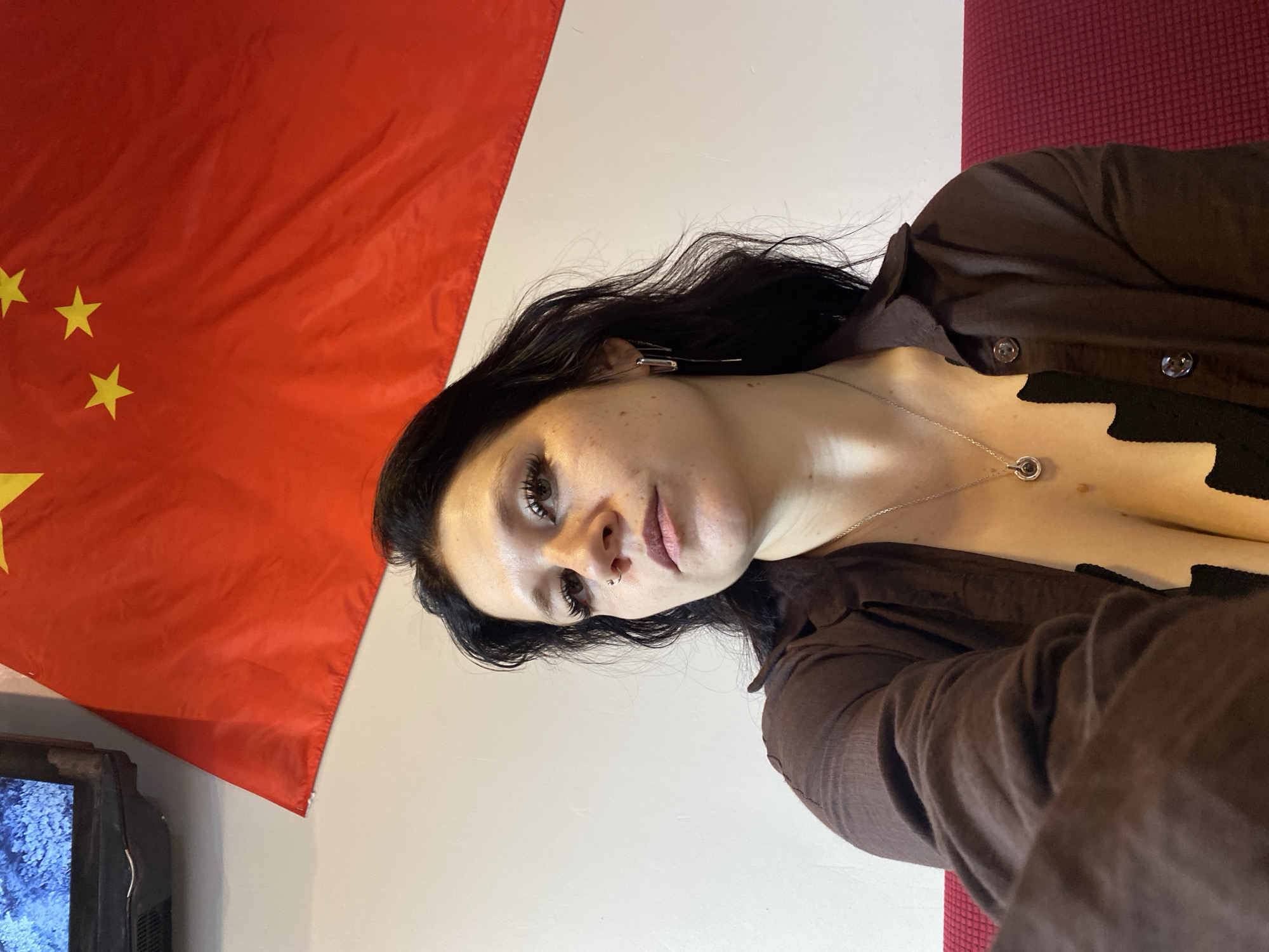 A selfie is taken by a woman with dark hair, wearing a brown shirt and a necklace. She has makeup on her eyes and nose ring. Behind her, a red flag with yellow stars is displayed, which appears to be the flag of China. To the left of the flag, a portion of a television screen is visible. The woman is seated on a red couch. The scene is set against a white wall. The specific location in Saint Petersburg, Russia cannot be determined from the image.