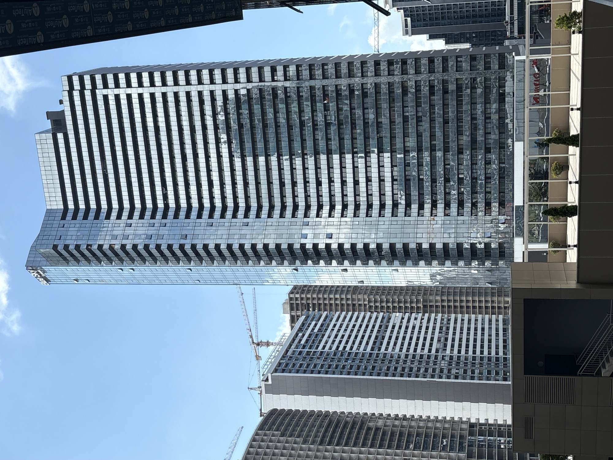 The image shows a cluster of modern high-rise buildings under a clear blue sky with scattered clouds. The central and tallest building is a sleek glass skyscraper with a prominent "Blue Sky" logo at its peak. Its facade is composed of numerous horizontal bands of windows and darker, reflective sections. To the left of this central tower, there are other buildings in various stages of construction, characterized by exposed concrete structures and scaffolding. One building on the far left is a gray, multi-story structure with visible grid-like patterns. Adjacent to the central skyscraper and slightly behind it is another tall, rectangular building with a facade of alternating dark and light horizontal bands, possibly indicating individual floors or sections. Further to the right, another glass-covered high-rise is visible, and beyond that, a shorter, dark-colored building. Several construction cranes are present in the background, suggesting ongoing development in the area. In the foreground, a portion of a rooftop or elevated walkway is visible with planters containing small trees and decorative lighting fixtures. The overall scene depicts a bustling urban environment with a focus on contemporary architecture and ongoing construction.