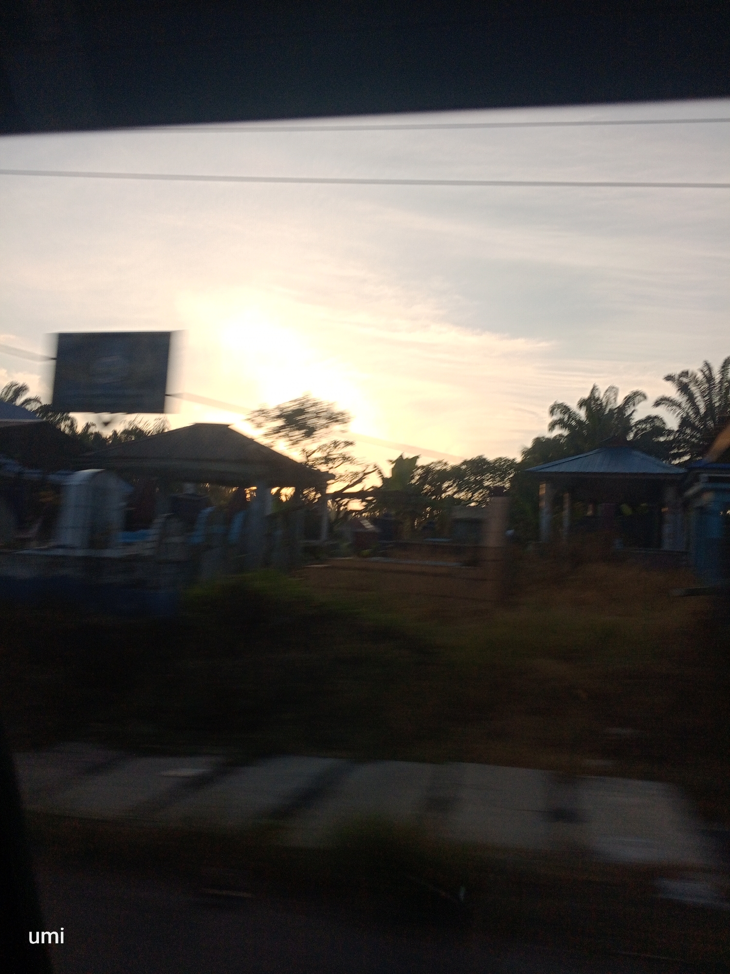 The image depicts a blurry outdoor scene at dusk, possibly in Bintang Meriah, Indonesia, with a faint sunset glow illuminating the sky. In the foreground, a dark, out-of-focus area obscures most of the ground, with the word "umi" visible in the bottom left corner.
The middle ground features a row of structures with dark roofs, appearing somewhat dilapidated. These structures are bordered by greenery, with palm trees and other foliage visible. One structure has a dark, angled roof and open sides, with faint shapes that could be people or objects within. A blurred black rectangular sign is positioned above and to the left of these structures.
The background is dominated by the hazy sky, with the sun low on the horizon, casting a soft, yellowish light. The overall image is characterized by motion blur and low light, making specific details difficult to discern.
