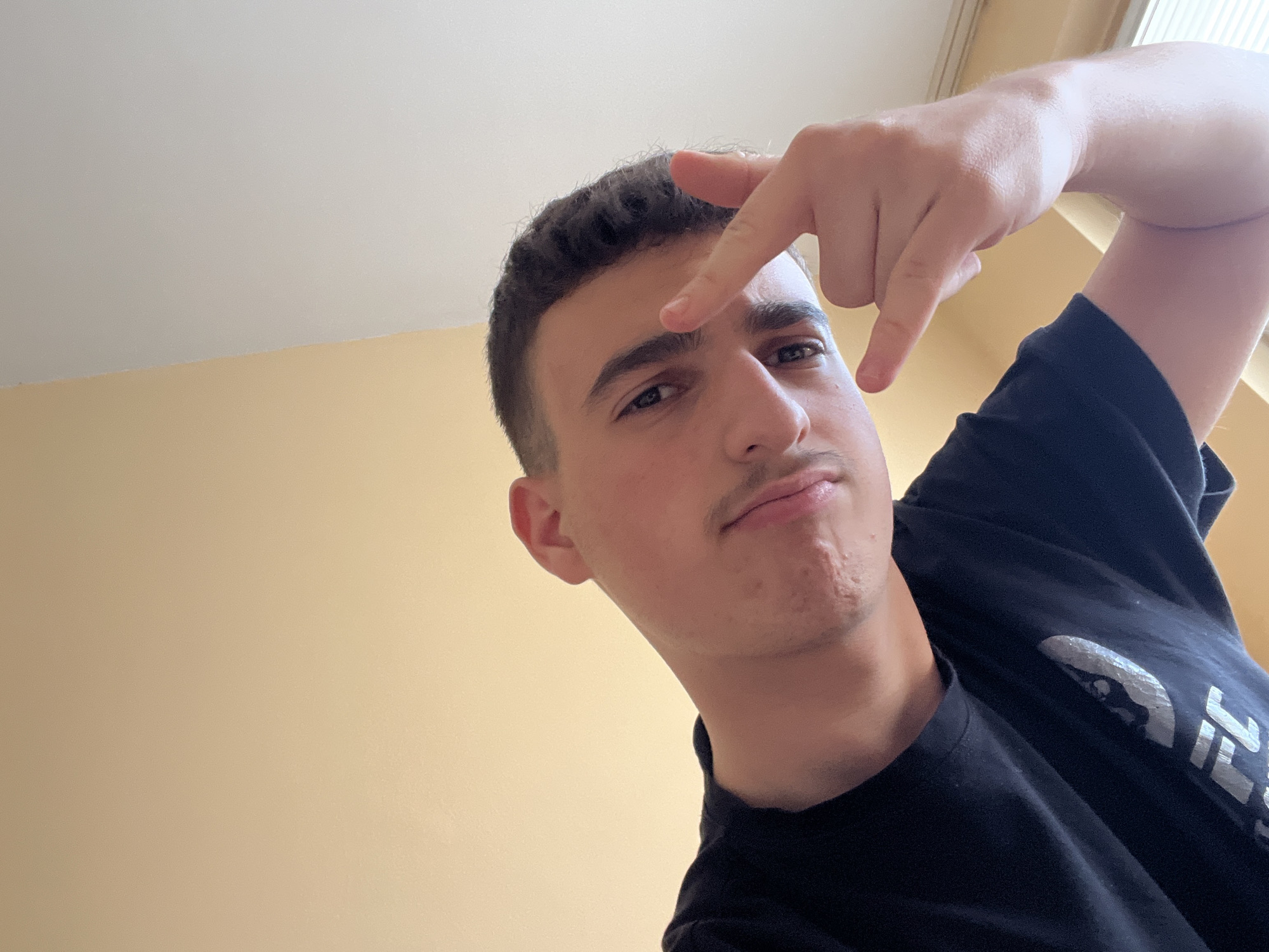 A light-skinned young man wearing a black t-shirt poses for a selfie. He has dark hair cut short and a thin mustache. He is making a hand gesture, pointing down towards his eye with his index finger. The background consists of a cream-colored wall and a white ceiling. The location is presumed to be indoors, possibly a room, but there are no specific landmarks or signs to confirm Kardzhali, Bulgaria.