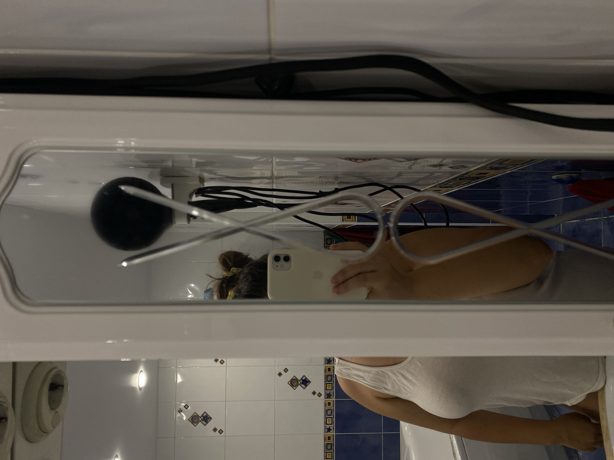 A person is taking a mirror selfie in a bathroom. The person is wearing a white tank top and has their hair up in a bun. They are holding a white smartphone in their right hand, which is being used to capture the reflection in the mirror. The bathroom has white tiled walls with a blue tiled border near the bottom. A bathtub is visible in the reflection on the left side of the image. On the right side of the mirror, black cables are visible, possibly connected to a hairdryer or other electrical appliance. The mirror itself has a decorative, ornate frame. The lighting suggests that the photo was taken indoors, likely in a residential bathroom. The image does not provide any specific visual cues that would allow for pinpointing the location to the city of Perm, Russia.