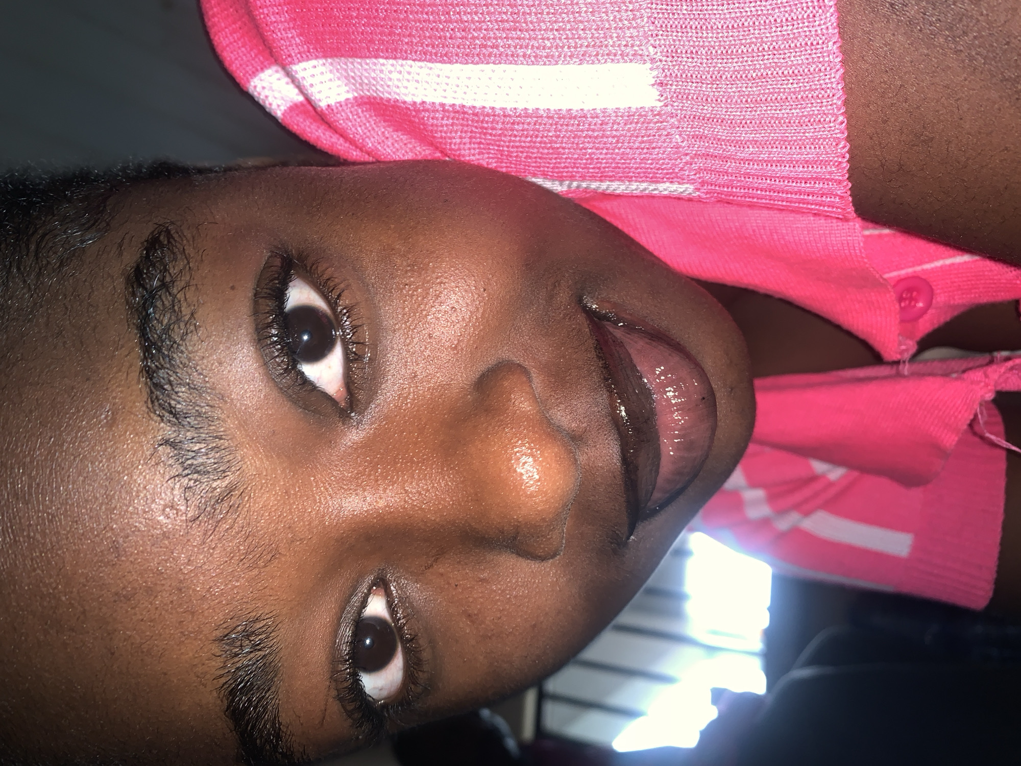 A close-up, low-angle shot captures the face of a young Black individual wearing a pink and white striped garment. The subject's eyes are dark and looking towards the camera, with visible eyelashes. Their mouth is slightly open, revealing a glistening tongue, suggesting an action or expression. The skin appears to have a natural sheen. The background is out of focus, with some bright lights visible on the left, possibly indicating an indoor setting with artificial lighting or a window. The image is dominated by the subject's face and upper body, with the texture of the fabric being a prominent element. No specific location in New Stadium, Nigeria, can be determined from this image.
