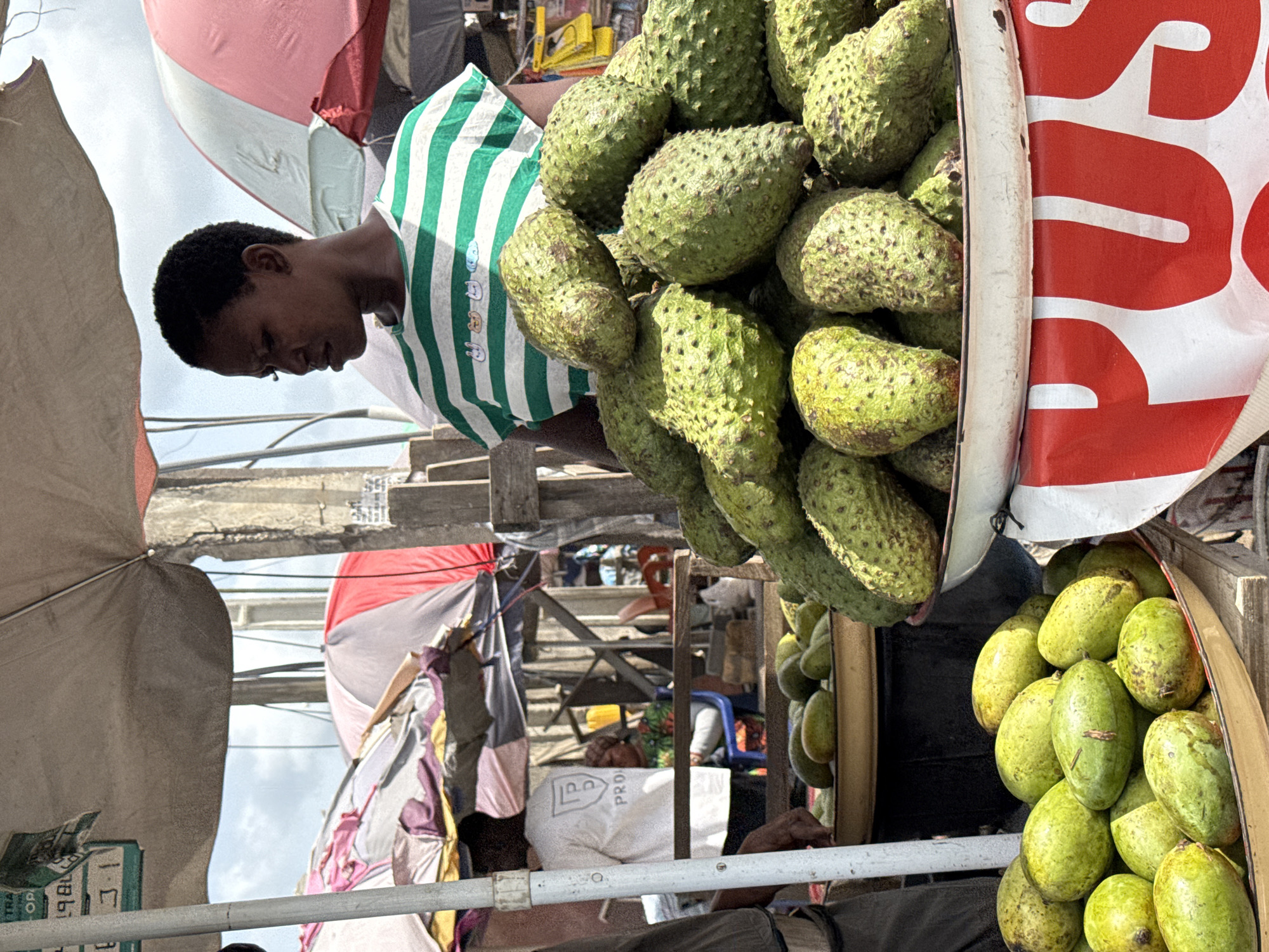 This image captures a vibrant market scene in Lekki, Nigeria, focusing on a fruit stall under a bright, cloudy sky. The central figure is a young person, seen in profile, wearing a green and white striped t-shirt. They are looking down, seemingly attending to the fruit display, suggesting they are a vendor or assistant.
The stall prominently features a large quantity of green, spiky soursop fruits, piled high in a white container on the left side of the frame. To the right, another container holds numerous smooth-skinned, green-yellow fruits, likely mangoes, indicating a diverse offering.
The market environment is characterized by an array of makeshift structures, including wooden supports and metal poles. Several umbrellas provide shade, with visible colors including red, pink, gray, and a patterned one with purple and yellow hues, hinting at a bustling outdoor setting. In the background, other market activities, partial figures, and more distant stalls can be discerned. A person is seated in the background, near a white bag with the partially visible text "PRO."
Visible text includes "RUS" in large white letters on a red background, which is part of the branding on the fruit container. Another small box or crate in the lower left shows numbers "1886" and letters "CO". The time of day appears to be daytime due to the bright light and visible sky. The overall impression is that of a lively and active local market.
