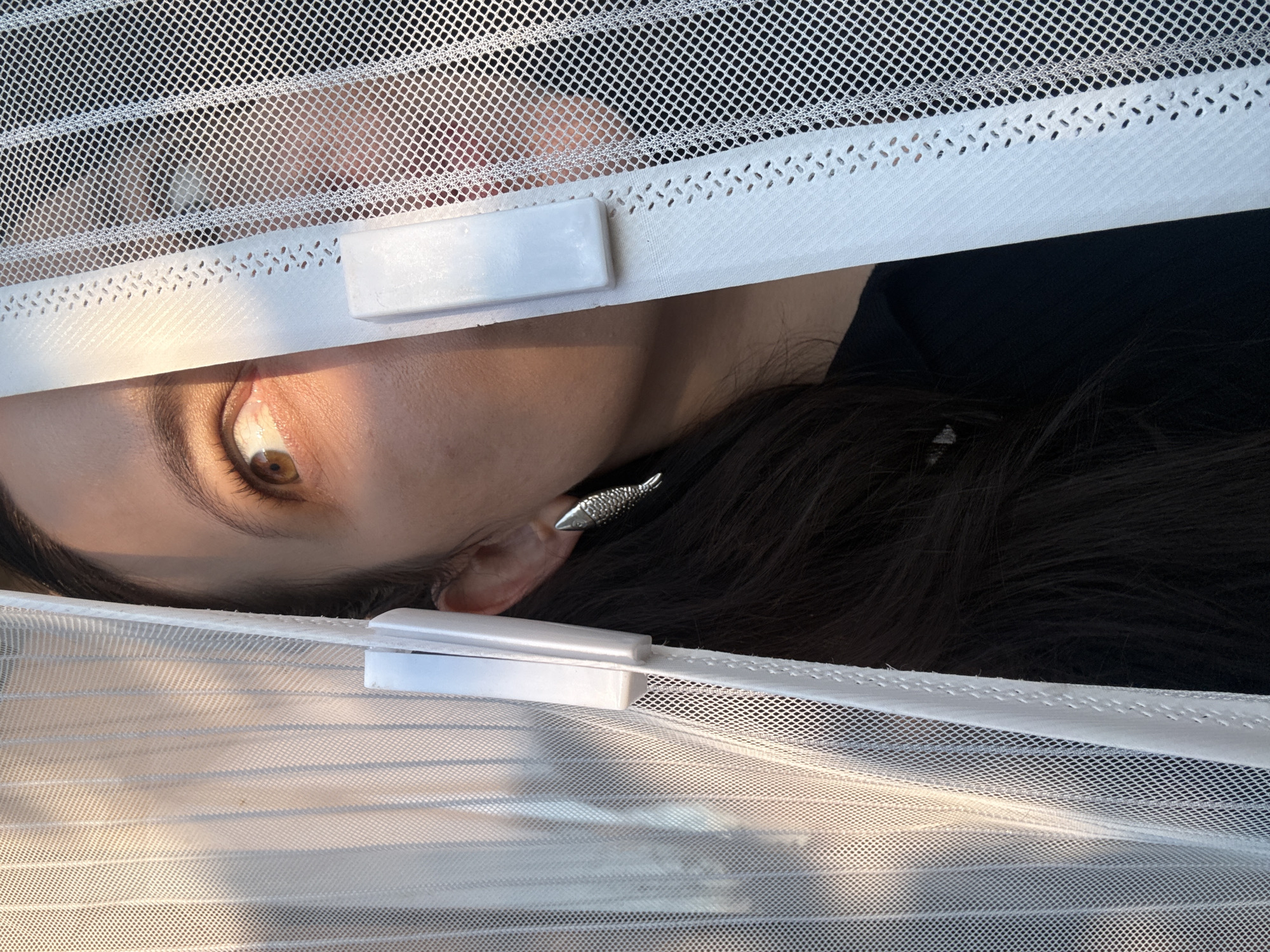 The image depicts a close-up of a woman partially obscured by a white mesh curtain. Her right eye, with a brown iris and dark eyelashes, is visible and catches the light from the left, casting a warm glow on her cheek. She is wearing a silver earring shaped like a fish. The curtain is held in place by two white fasteners. The background appears to be outdoors, with diffused light suggesting either early morning or late afternoon sun. The woman's dark hair is also visible, framing her face. The scene is likely a residential setting in Chaikovsky, Russia, given the context provided.