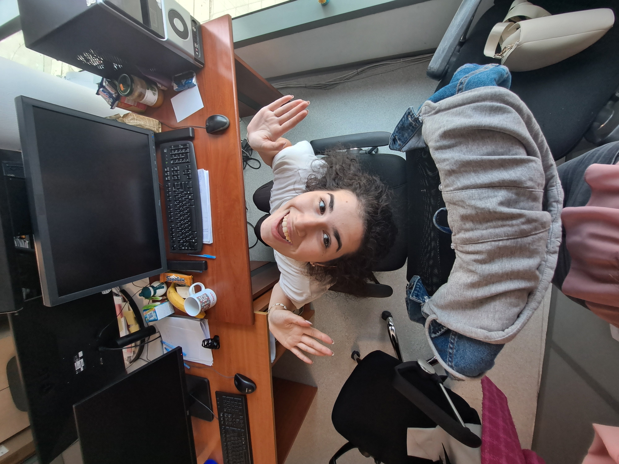 A high-angle, rotated shot captures a young woman smiling brightly at the camera, her hands raised in a welcoming gesture. She has curly dark hair and wears a white t-shirt. The setting appears to be an office or study environment, seen from a perspective looking down onto a cluttered wooden desk and the surrounding area.
The woman is seated on a black office chair. To her left, the wooden desk holds multiple computer monitors, two black keyboards, and computer mice. Various items are spread across the desk, including a white mug with the visible text "MAMY" and a pattern, a yellow banana, a jar of what appears to be preserves or food, a golden-wrapped snack with "Tropileu" visible, and stacks of papers and pens. A desktop computer tower is mounted under the desk's edge.
Beyond the woman, another black office chair holds a grey hooded sweatshirt and blue jeans, with a light-colored shoulder bag or purse resting on it. The floor is light-colored, and in the background, cables are visible running along a wall or partition, suggesting a typical office setup. The overall mood is cheerful and informal, with the woman's joyful expression dominating the frame.