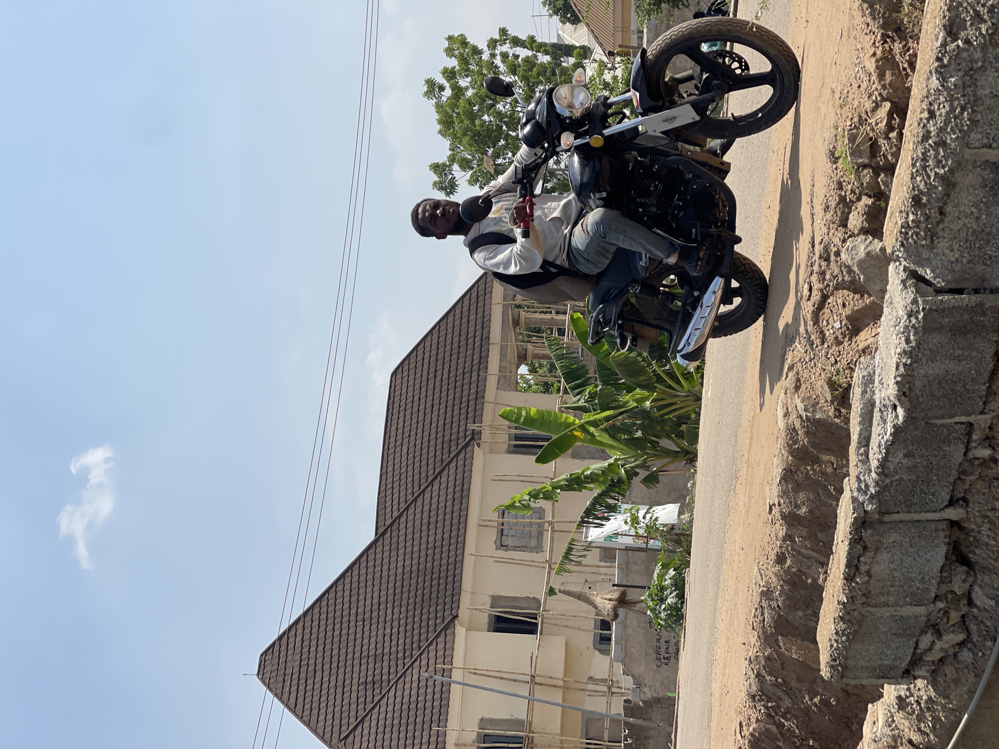 A young man is prominently featured, seated on a black motorcycle on an unpaved road under a clear blue sky, in what appears to be a residential area of Abuja, Nigeria. The image is taken outdoors in bright daylight, suggesting midday or early afternoon.
The man, with dark skin and a short beard, wears a light-colored long-sleeved top and light blue jeans. He looks directly at the camera with a neutral expression, his hands on the motorcycle's handlebars, one of which has a distinctive red grip. The motorcycle is a black model, possibly a cruiser, with chrome accents on its exhaust pipe and other parts. The word "CRUISER" is faintly visible on a silver plate near the rear wheel.
In the background, a two-story building with a dark brown tiled hip roof stands partially constructed or renovated, evidenced by visible bamboo scaffolding on its light-colored (cream or pale yellow) walls. Several dark window openings are visible. Lush green trees and large-leafed plants, possibly banana trees, surround the building. The road beneath the motorcycle is dirt or gravel, and to the immediate right, a raised bank of rough soil and scattered concrete blocks marks the edge of the road or an unfinished area. Thin power lines are visible against the sky in the upper right. Partially visible text on a wall in the background reads "GENERAL REPAIR".