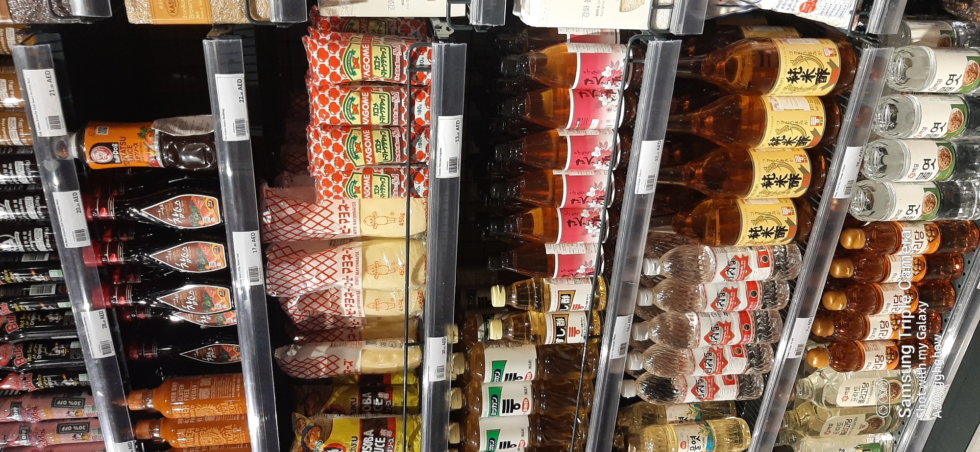 The image shows a shelf of assorted condiments and sauces in what appears to be a supermarket aisle. The products are arranged on multiple shelves, with many bottles and packages visible. The lighting suggests an indoor retail environment. There are no people visible in the image. The product labels are primarily in Asian languages, suggesting an international or Asian specialty store. The price tags visible indicate prices in AED (United Arab Emirates Dirham), and the presence of "Dubai, United Arab Emirates" in the prompt confirms the location. The items include bottles of what seem to be soy sauce or similar dark liquids, mayonnaise packets, ketchup packets, and other bottled sauces and vinegars. The overall impression is of a well-stocked retail display of food products.