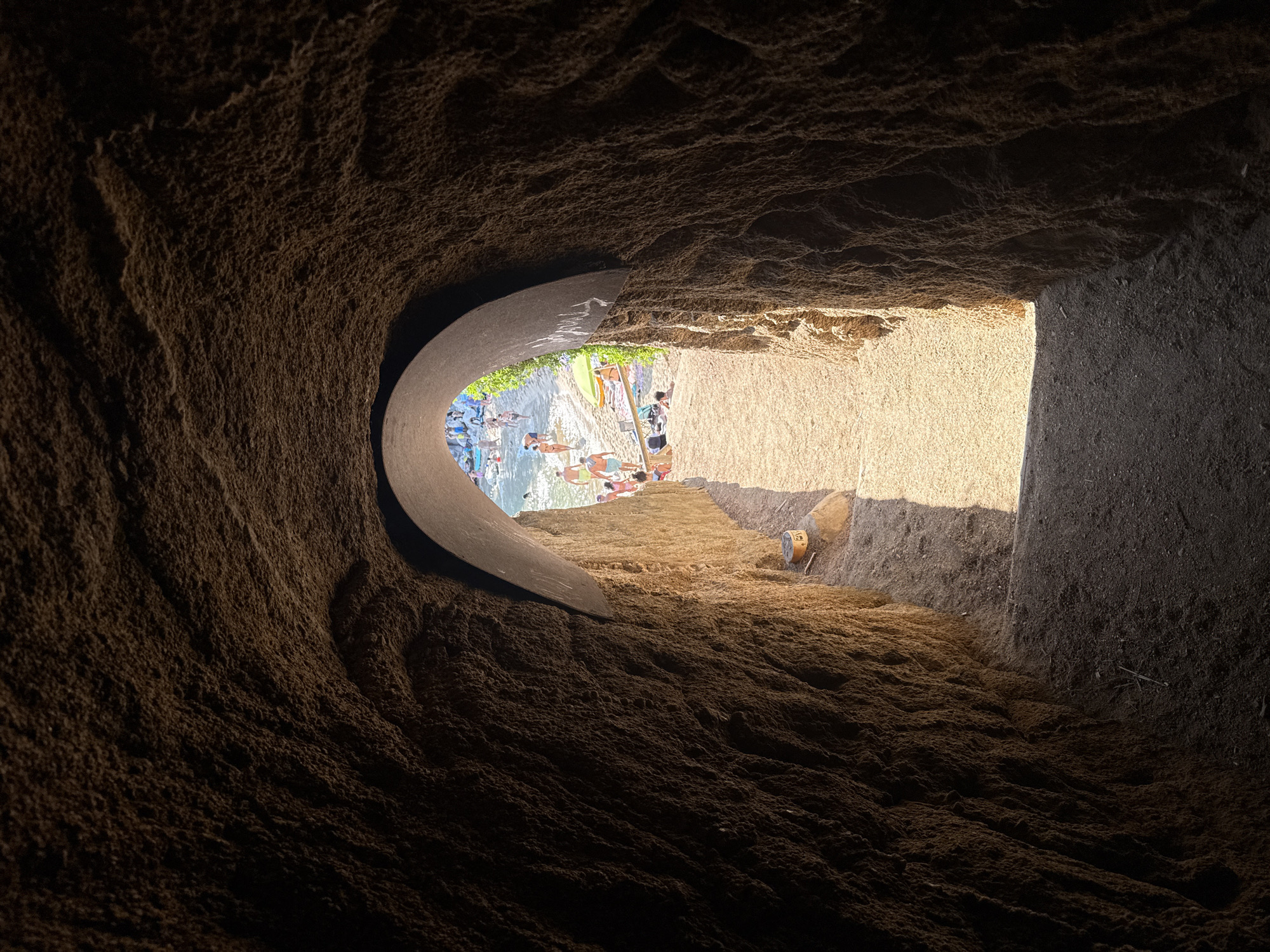 This image shows the interior of a natural rock tunnel opening onto a crowded beach in Platja d'Aro, Spain. The tunnel walls are rough and textured, appearing to be made of a sandy, reddish-brown rock. Sunlight streams in from the opening, illuminating the sandy path leading out of the tunnel and the beach beyond.
On the beach, numerous people are visible, some wading in the water, others sunbathing on towels or under umbrellas. There are lounge chairs and beach umbrellas scattered across the sand. The water appears calm and clear. The scene suggests a popular and vibrant beach destination.
The lighting within the tunnel is dim, with a distinct beam of light cutting across the path and the tunnel floor, suggesting the sun is high in the sky. The contrast between the dark interior of the tunnel and the bright, sunlit beach creates a dramatic effect. A small, cylindrical object with markings is visible on a rock to the right of the tunnel exit.
