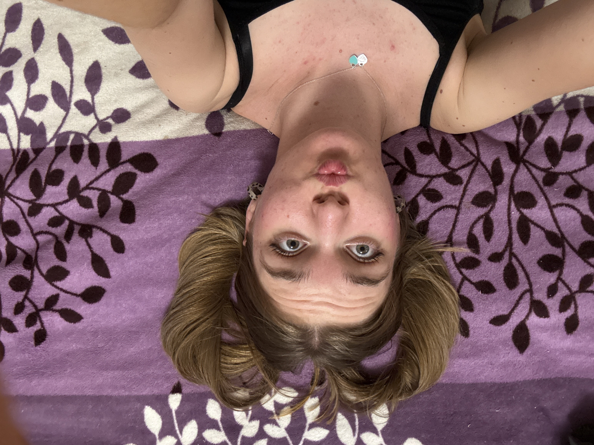 A woman is lying on a blanket with a pattern of white and brown leaves on a purple background. She is wearing a black top and a silver necklace with a blue charm. Her blonde hair is in pigtails. The photo is taken from an overhead perspective. The location cannot be determined from the image.