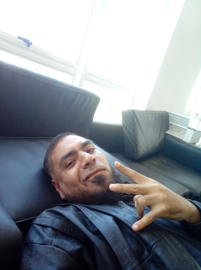 An indoor scene depicts a male individual reclining on a dark-colored sofa. The subject is positioned facing the camera, with a slight smile and direct gaze. He has dark hair, a trimmed beard in a goatee style, and an apparent fade haircut on the sides. He is wearing a dark, possibly leather, jacket or shirt. His left hand is raised, forming a "peace" sign (index and middle fingers extended in a 'V' shape). The background features large white-framed windows emitting significant natural light, causing overexposure in parts of the upper frame. To the upper right, a white object resembling a printer or scanner is visible on a surface against a light wall. The specific location within the city of Guatire, Venezuela, cannot be determined from the visual information provided.