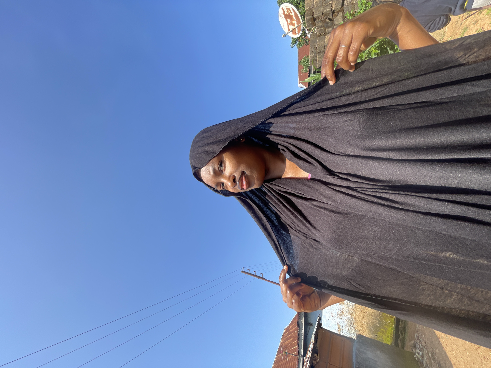 A Black woman is pictured outdoors under a clear blue sky, wearing a black hijab that drapes over her shoulders. Her left hand is holding the hijab up near her face, and her right hand is adjusting it near her shoulder. In the background, to the left, there is a building with a rusted corrugated metal roof, a utility pole with wires, and a concrete structure. To the right, there is a brick wall with a satellite dish mounted on it and some green foliage. The ground appears to be dirt. This scene is consistent with residential areas in cities like Jos, Nigeria, where such structures and elements are commonly found.
