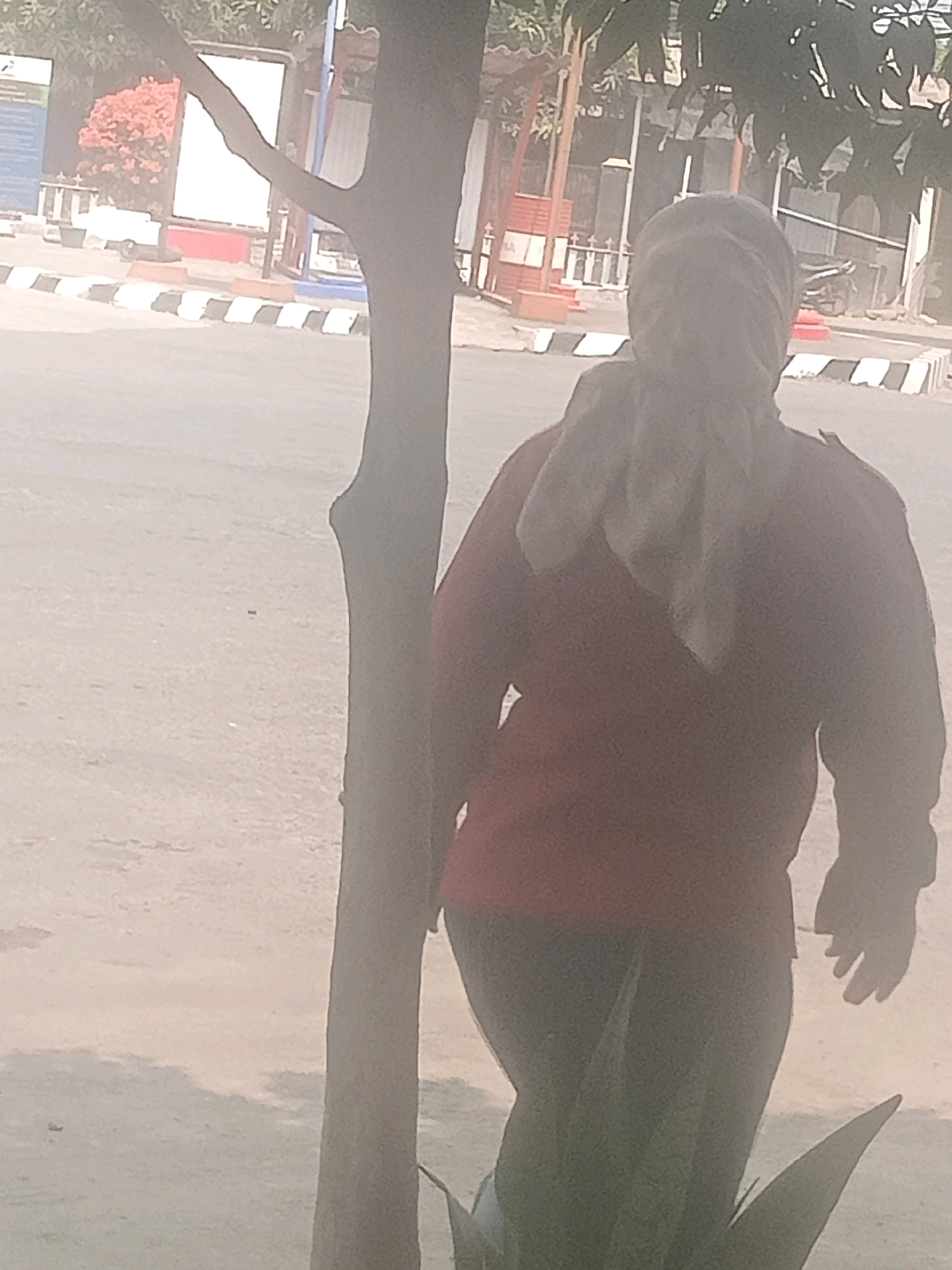 The image shows a person from behind, wearing a red long-sleeved shirt and dark pants. Their head is covered by a light-colored scarf, obscuring their face. The person appears to be walking or standing near a tree trunk, which is prominently positioned in the foreground. In the background, there is a street with a checkered black and white border, suggesting it might be a pedestrian crossing or a designated area. Beyond the street, structures that resemble buildings or stalls are visible, some with red accents. The environment seems to be outdoors, possibly in a public area within the city of Pendawa, Indonesia. The overall lighting suggests it is daytime, with a slightly hazy atmosphere.