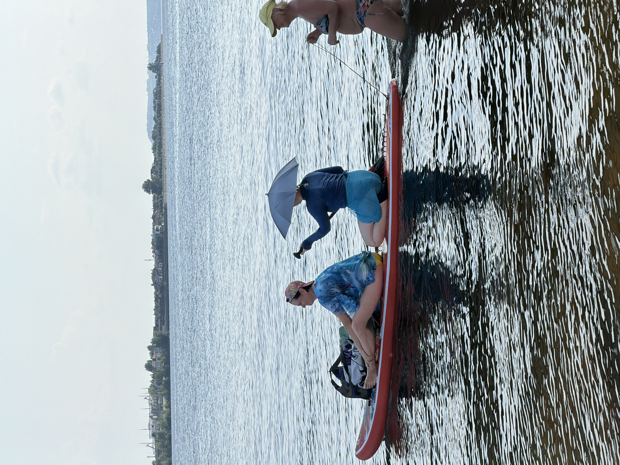 Three individuals are present on the water. The first individual, seated on a red paddleboard, wears a blue tie-dye shirt and yellow shorts, with a pink and white patterned bandana on their head. They are accompanied by a large duffel bag. The second individual is also seated on the paddleboard, wearing a blue long-sleeved shirt and blue shorts, holding a gray umbrella over themselves. The third individual, partially submerged in the water, wears a yellow cap and a patterned bikini, pulling the paddleboard with a rope. The paddleboard is on a body of water with a shoreline visible in the background, featuring buildings, trees, and distant hills under a cloudy sky. The scene appears to be on the Volga River in Saratov, Russia.