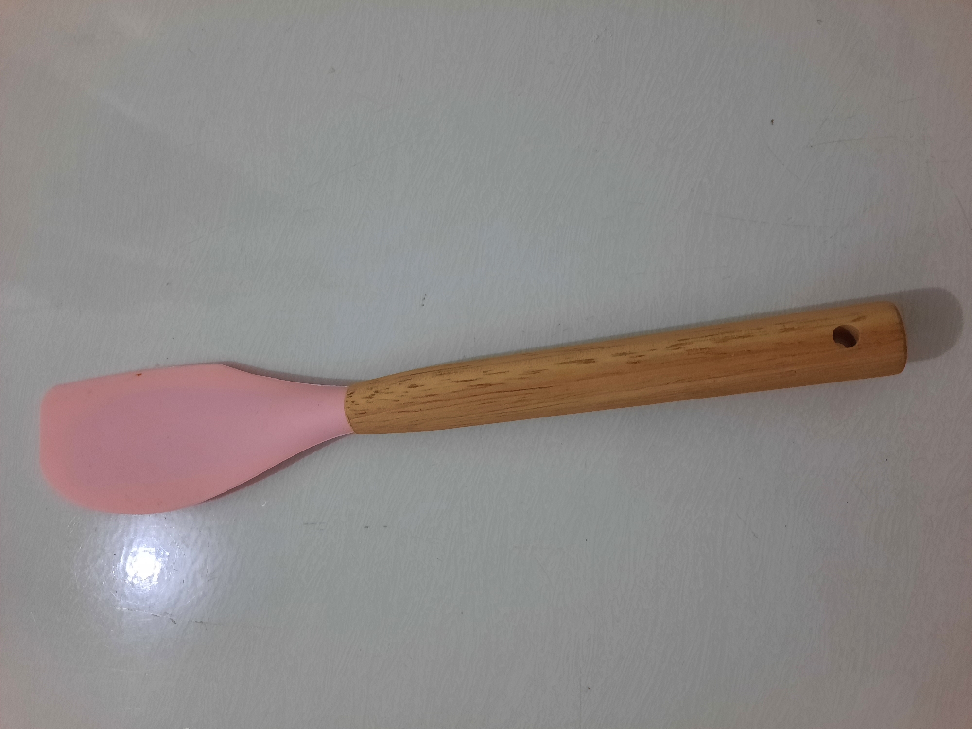 A single pink silicone spatula with a wooden handle is positioned diagonally on a textured, light-colored surface. The spatula is oriented with the handle towards the bottom right and the silicone head towards the top left. The surface appears to be a countertop or tabletop, exhibiting subtle linear patterns and some minor scuffs. The lighting creates a slight reflection on the surface, primarily near the top left quadrant. The image is a close-up view of the object, with no other items or people visible. The background provides no specific geographic context for Lhokseumawe, Indonesia, other than the possibility of it being an indoor domestic or commercial setting within the city.