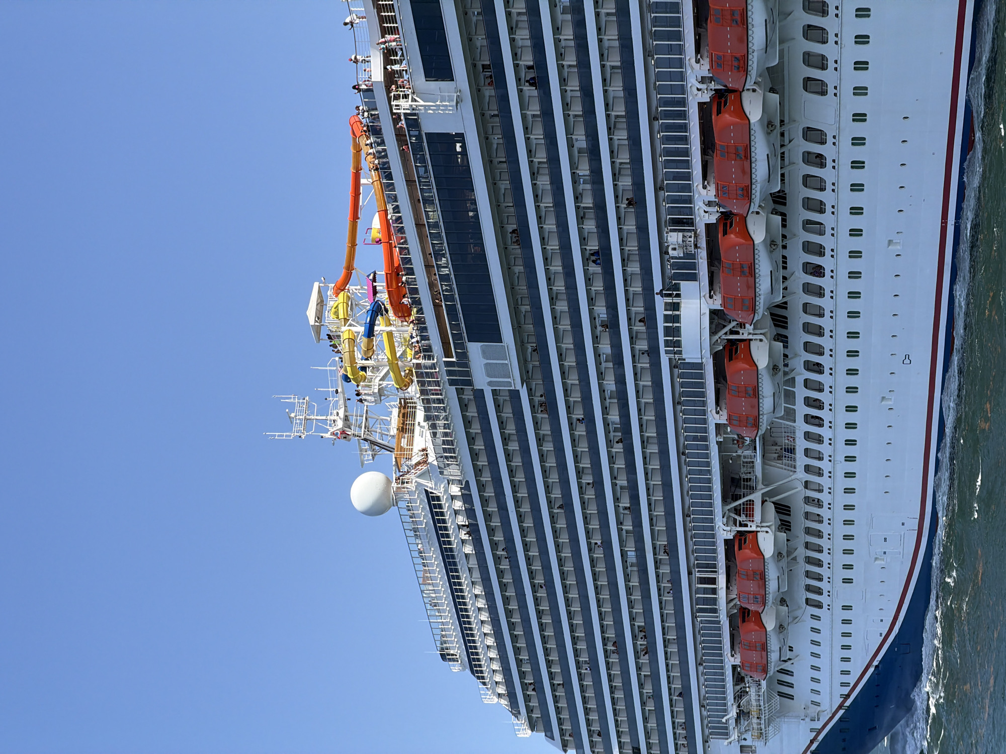 A large white cruise ship is sailing on the water under a clear blue sky. The ship is angled from the left to the right of the frame, with its port side prominently displayed. Multiple decks of cabins with dark-rimmed windows are visible. A row of bright orange lifeboats is attached to the side of the ship, above a lower deck with numerous small windows. At the top of the ship, colorful water slides are visible, along with several people standing on decks and walkways. The water around the ship is choppy, indicating movement. The image appears to have been taken during daylight. The ship is not in Miami Beach, but on the open water.