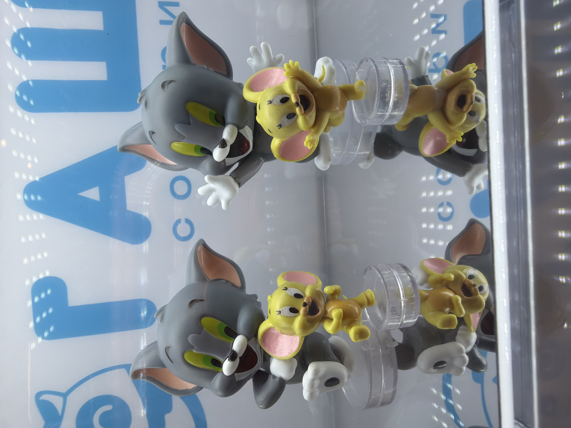 The image shows two cartoon character figurines of Tom and Jerry inside a glass-fronted vending machine. Tom, a gray cat, is depicted with a slightly menacing expression. Jerry, a yellow mouse, is clinging to Tom's head. The figurines are attached to a clear plastic structure with white detailing. The background is a blurry white surface with blue lettering. The scene appears to be indoors due to the vending machine.