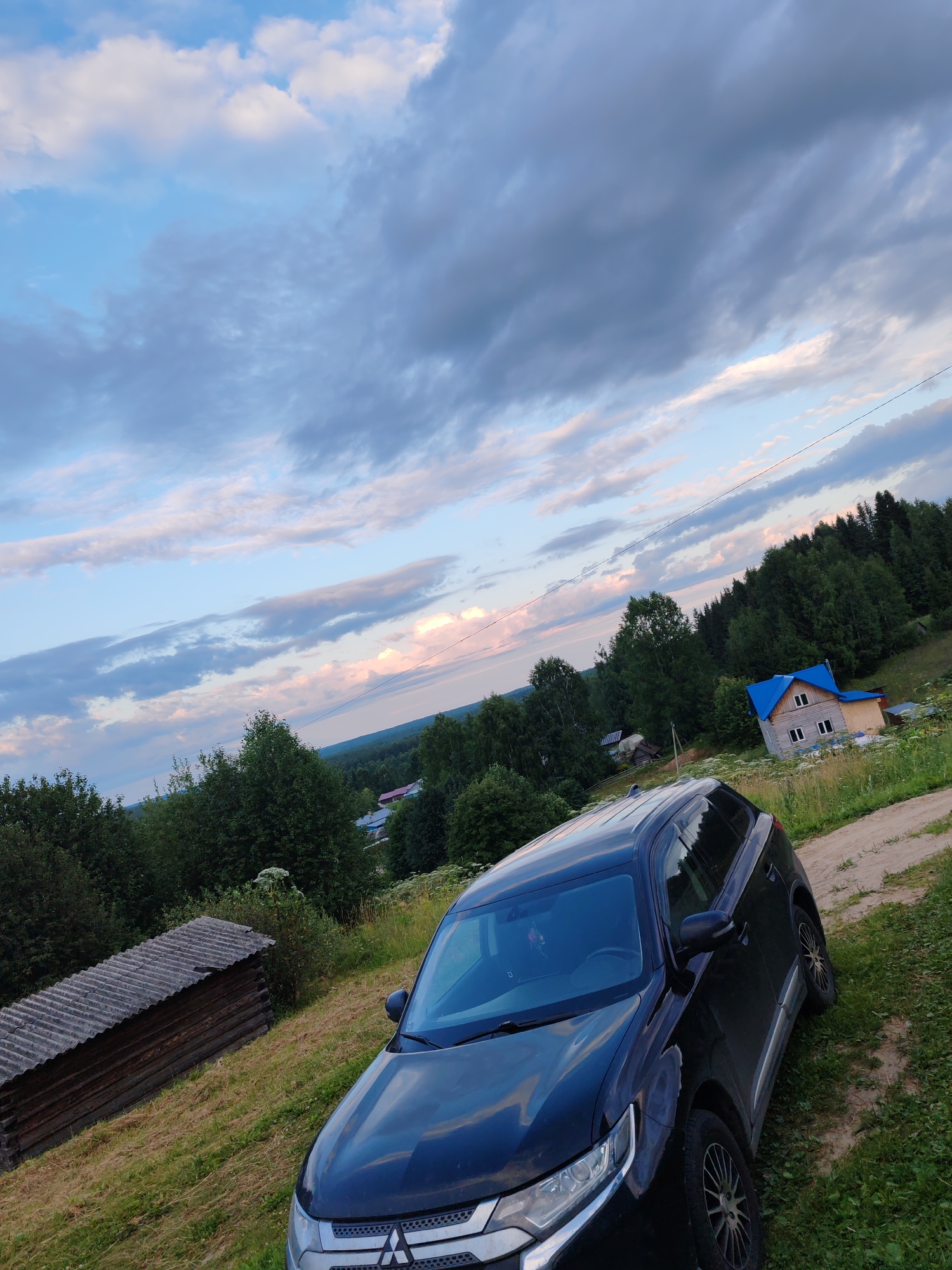 The image captures a rural scene with a black SUV parked on a dirt path in what appears to be the city of Chukhloma, Russia. The SUV is angled towards the right side of the frame. In the background, there is a wooden structure with a corrugated roof, and beyond that, a forest covers a hillside. A house with a blue roof is situated further up the hill. The sky above is partly cloudy with streaks of pink and blue hues, suggesting either sunrise or sunset. The ground is covered in grass, and a dirt road leads away from the SUV and into the distance.