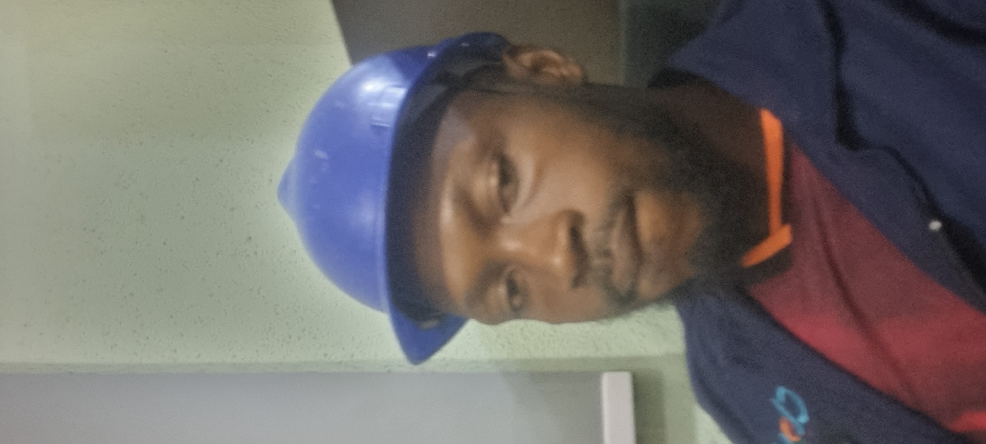 In Ikeja, Nigeria, a close-up, slightly blurry photograph shows a Black man wearing a blue hard hat and a dark blue jacket over a red shirt. The man has a beard and appears to be looking down and to his left. The background is a textured, pale green wall.