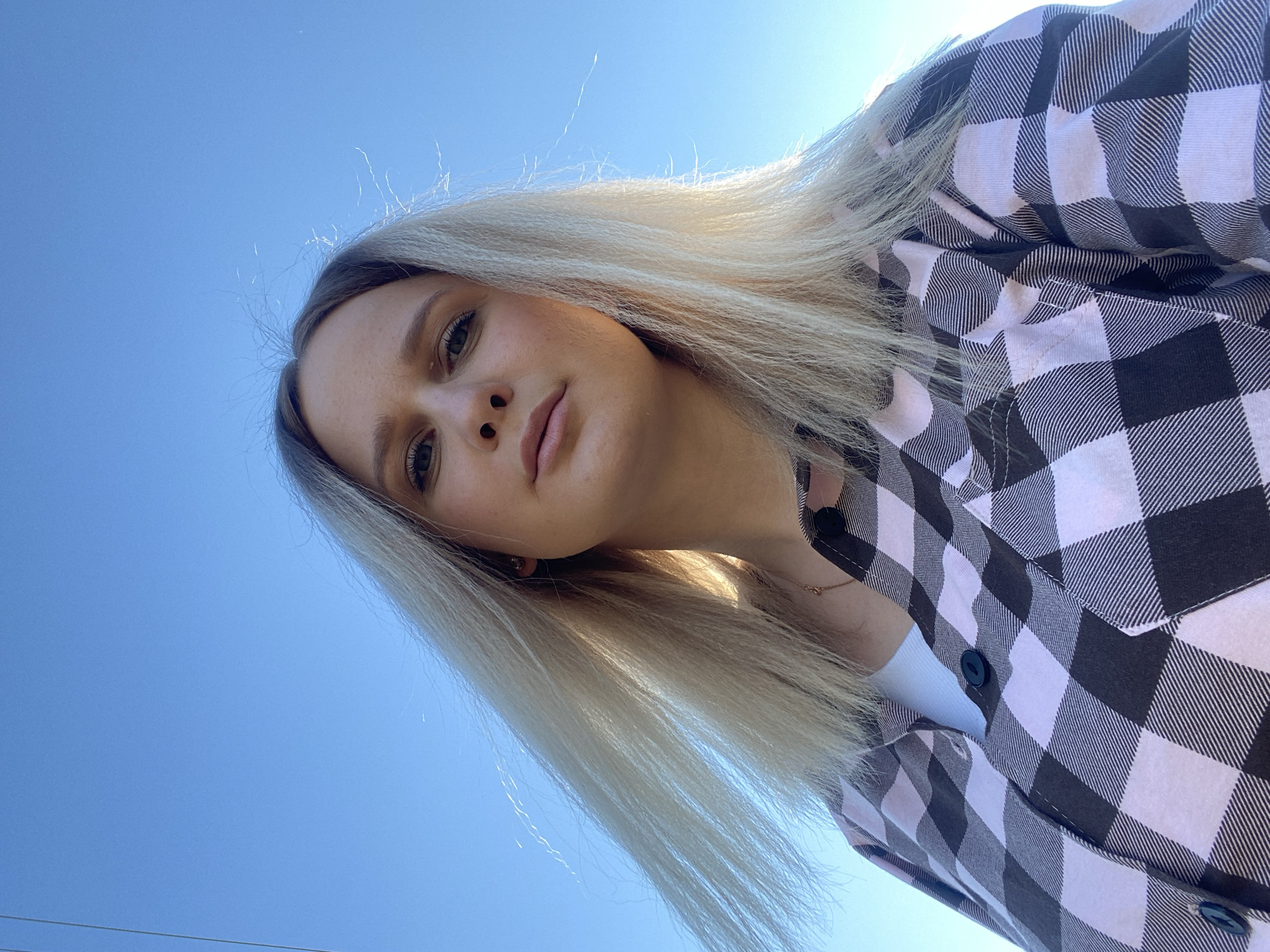 A young woman with long, straight blonde hair is featured in a brightly lit, upward-angled selfie. The image is tilted diagonally, with the woman's head occupying the upper-middle portion. She has a neutral expression, looking directly into the camera with her eyes. Her fair skin is softly illuminated by natural daylight, highlighting her features.
She is wearing a button-up shirt with a black and white (or very pale pink and black) plaid pattern, worn over a white undershirt. A delicate gold chain necklace is visible around her neck.
The background is a clear, vibrant blue sky, indicating an outdoor setting on a sunny day. Sunlight catches on the fine strands of her hair, creating a luminous effect against the bright sky. The perspective suggests the camera was held low, looking up at the subject. The scene appears to be set outdoors in bright conditions, possibly in Kauchuk Stantsiya, Russia. There is no visible text in the image.