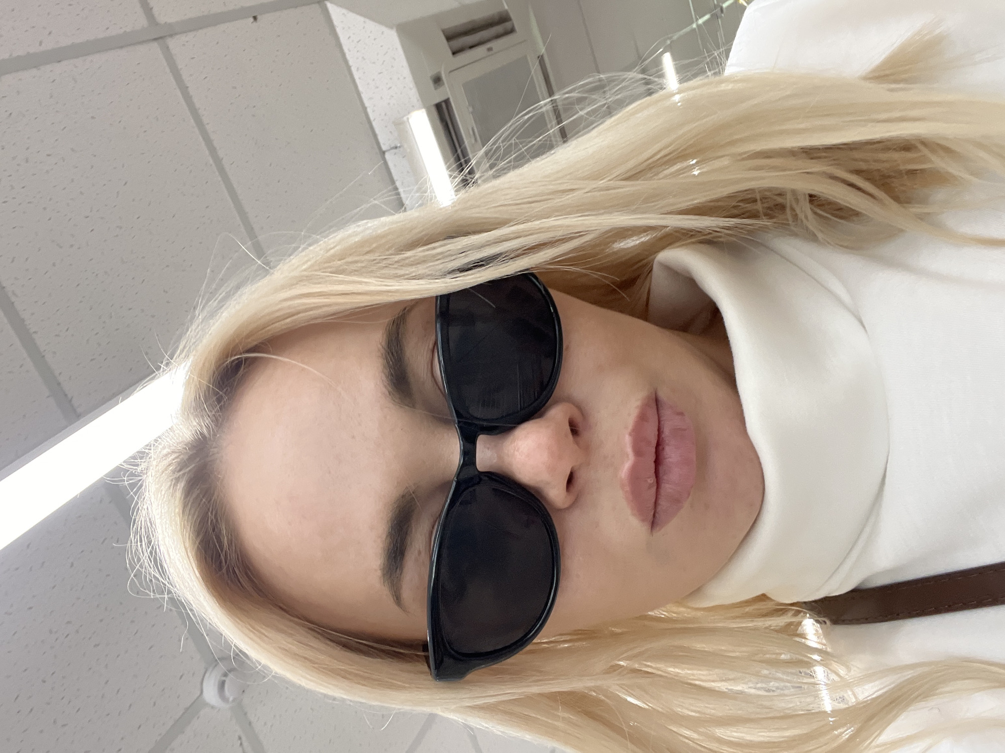 A close-up, high-angle selfie of a blonde woman wearing black sunglasses and a white turtleneck sweater. Her lips are slightly parted, and her hair is styled loosely around her face. The background appears to be an indoor environment with a tiled ceiling and fluorescent lighting. There is a brown strap visible on her shoulder. The woman is positioned in the center of the frame, with her face taking up most of the image.