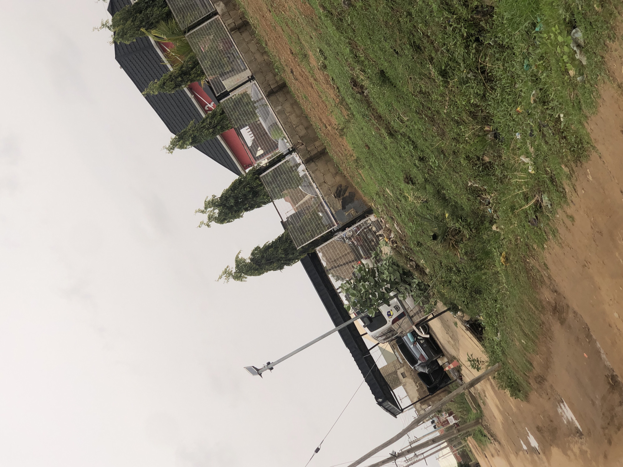 The scene depicts a roadside view in Hanwa, Nigeria. The sky is overcast. A row of buildings with black roofs and wire mesh enclosures lines the left side of the image. A light pole is visible in front of one of the buildings. A grassy area and a dirt road occupy the right side of the frame. There are no people immediately visible, though cars can be seen by the buildings. Power lines are visible. Debris and trash are scattered throughout the greenery.