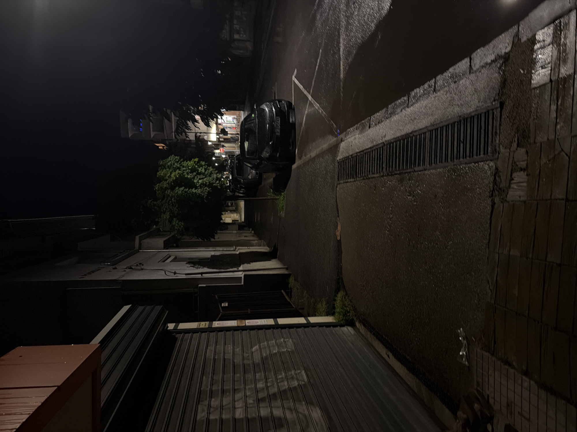 This media file captures a nighttime street scene in Pirgovo, Bulgaria, likely following rainfall or in very damp conditions, indicated by the wet and reflective surfaces. The image is oriented vertically (rotated 90 degrees clockwise from a typical street view), showing the ground plane and buildings extending upwards.
No people are visible in the static scene.
In the mid-ground, a dark, wet asphalt street stretches horizontally across the frame (when correctly oriented). Faint white road markings are visible. A black SUV is parked on the left side of the street, facing left. Its license plate is partially visible but unreadable due to darkness and resolution. Adjacent to the right side of the street is a long, dark drainage grate embedded in a concrete structure, with the wet, textured wall rising behind it.
Buildings line both sides of the street. On the left side, beyond the SUV and interspersed with dark foliage, are multi-story buildings, some showing faint interior lights with cool blue and white hues. On the right, the immediate foreground features various dark-colored corrugated metal roofs or covers, along with a reddish-brown structure, suggesting garages or commercial units at street level. More dark building outlines are visible further down the street. The sky is completely dark, blending with the shadows of distant structures. The overall mood is quiet and subdued. No discernible text is clearly readable apart from the unidentifiable characters on the vehicle's license plate.