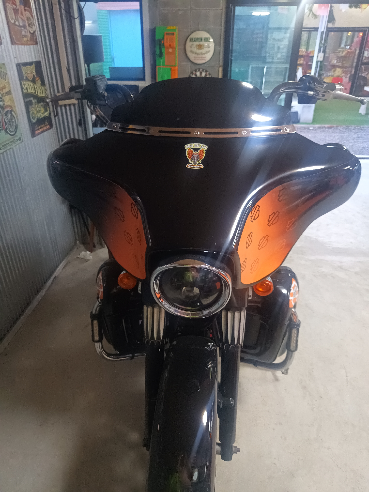 A black motorcycle with orange accents is parked in a garage in Surat Thani City Municipality, Thailand. The motorcycle features a large fairing with a logo and chrome accents. The surroundings include metal walls, windows, and other indoor garage elements.