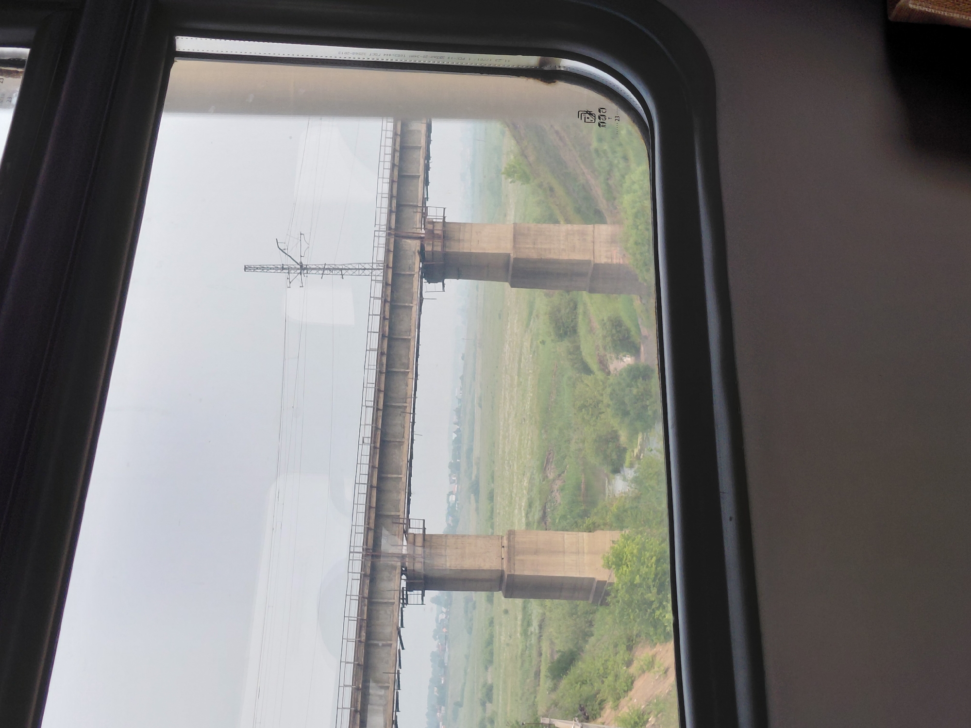 The image is a view from inside a train, looking out of a window at a concrete bridge spanning a grassy valley under an overcast sky. The bridge is supported by large concrete pillars. In the distance, a few buildings are visible, suggesting a rural or semi-rural setting. There are no people or animals visible in the frame. The scene is calm and static. The question about the location in Chik, Russia, cannot be answered as there is no information within the image to support this claim.