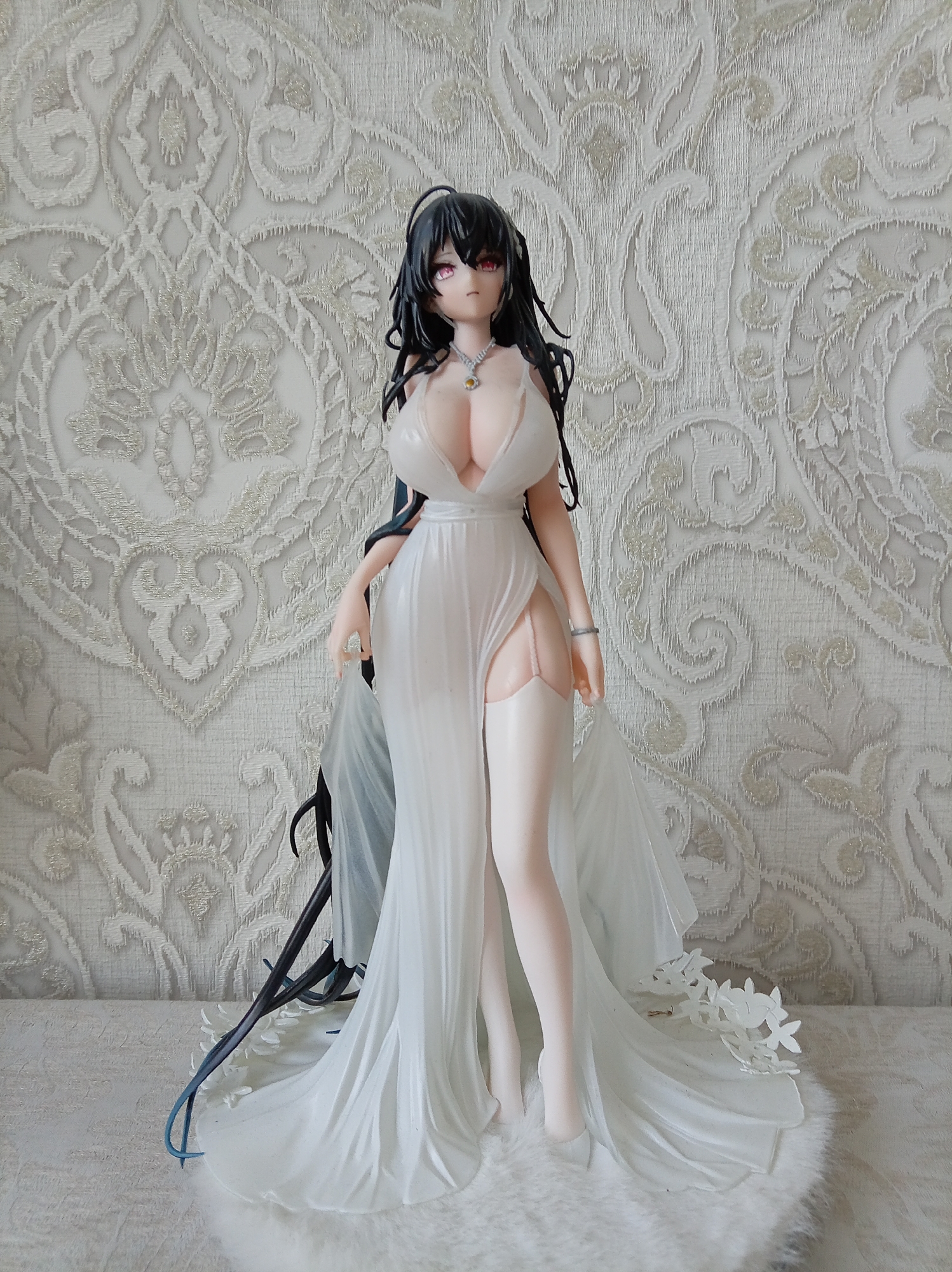 This is a full-body, eye-level shot of a meticulously detailed anime-style figurine of a young woman. She stands on a white, fluffy surface, likely a rug or faux fur, against a backdrop of ornate, light-colored textured wallpaper. The figurine depicts a character with long, black hair, striking red eyes, and pale skin. She is wearing a flowing, low-cut white dress with a high slit, revealing her legs and delicate stockings. A silver bracelet adorns her left wrist, and a necklace with a yellow pendant rests on her chest. The dress has sheer white fabric accents that trail behind her, adding to its ethereal quality. The figurine is the sole subject of the image, and there are no discernible environmental features that suggest a specific city or location like Yessentuki, Russia. The lighting is even, highlighting the intricate details of the figurine and the texture of the wallpaper.
