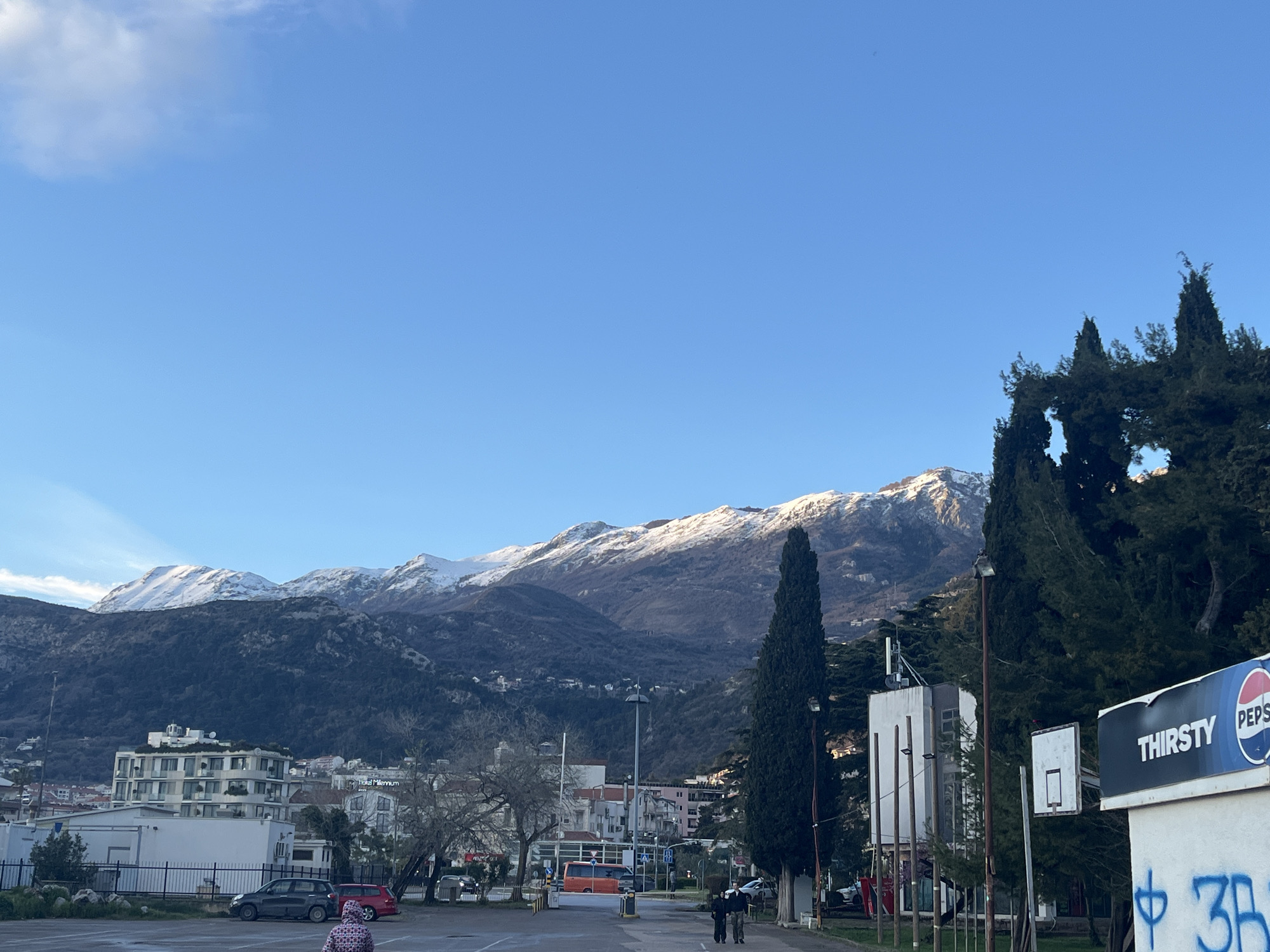 The image captures a street scene in Budva, Montenegro. Snow-capped mountains form the backdrop to the urban landscape. Buildings, trees, and parked cars are visible. Two pedestrians are walking along the street. A Pepsi sign is prominent in the foreground, along with a basketball hoop. Graffiti is present on the wall beneath the Pepsi sign. The sky is mostly clear blue with a few scattered clouds. The lighting suggests it is daytime.