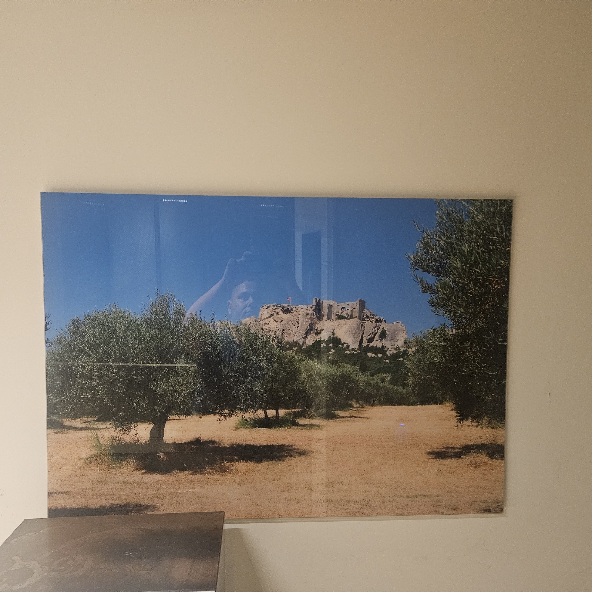 The image displays a framed photograph hanging on a wall. The photograph depicts a rocky outcrop with a ruined castle on top, set against a clear blue sky. In the foreground, there are several olive trees with dense green foliage. The ground beneath the trees appears to be dry, sun-baked earth with sparse grass. There is a reflection of a person, possibly the photographer, in the glass of the frame, with their hand reaching up towards the top of their head. The castle appears to be the Château de Lançon in Lançon-Provence, France, a historical landmark situated on a prominent rock formation.